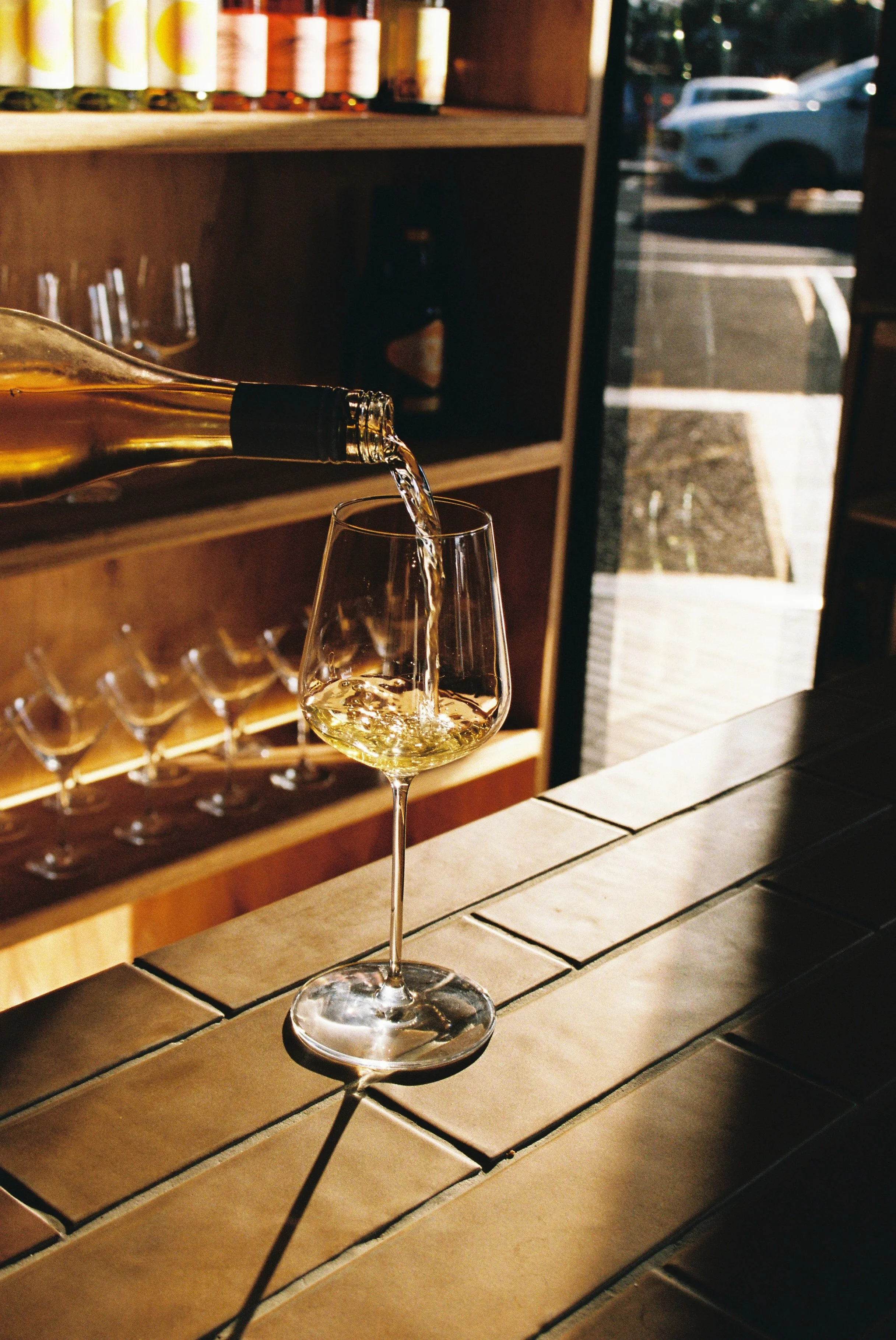 A wine glass on a tiled bar counter being filled with white wine from a bottle. In the background, a shelf with bottles and glasses can be seen near a window with sunlight and parked cars outside. Gold Coast Food Photography by Joey Bailey