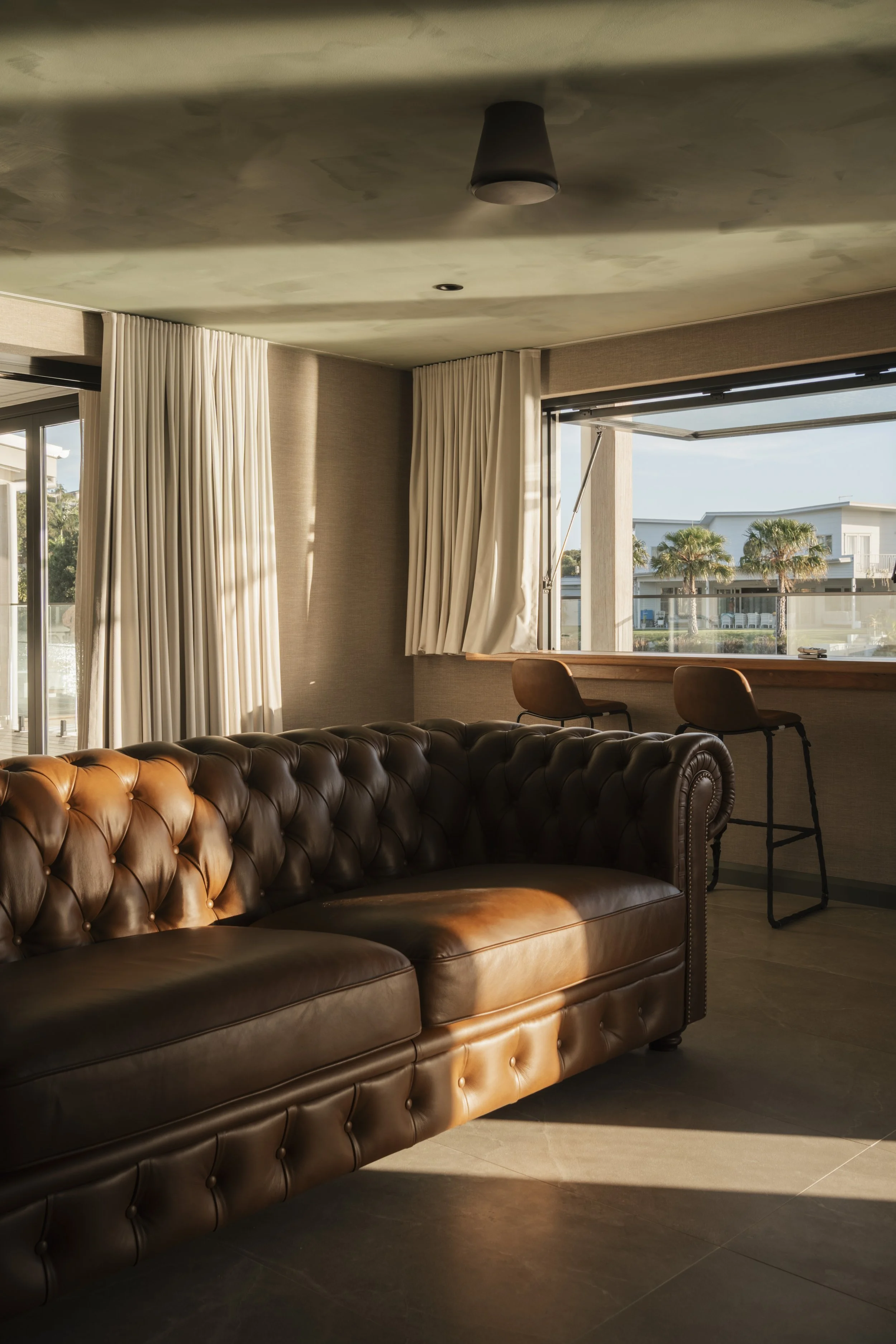 Sunlight streaming into a modern living room with a brown leather tufted sofa, white curtains, and a large window showing palm trees outside. Gold Coast Architecture photography Joey Bailey