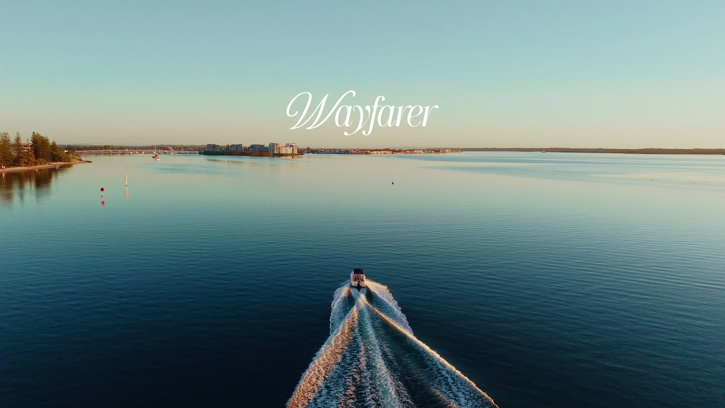 A boat speeds across calm water, creating a wake, with a skyline of buildings and trees in the distance under a clear sky. Gold Coast Construction videography
