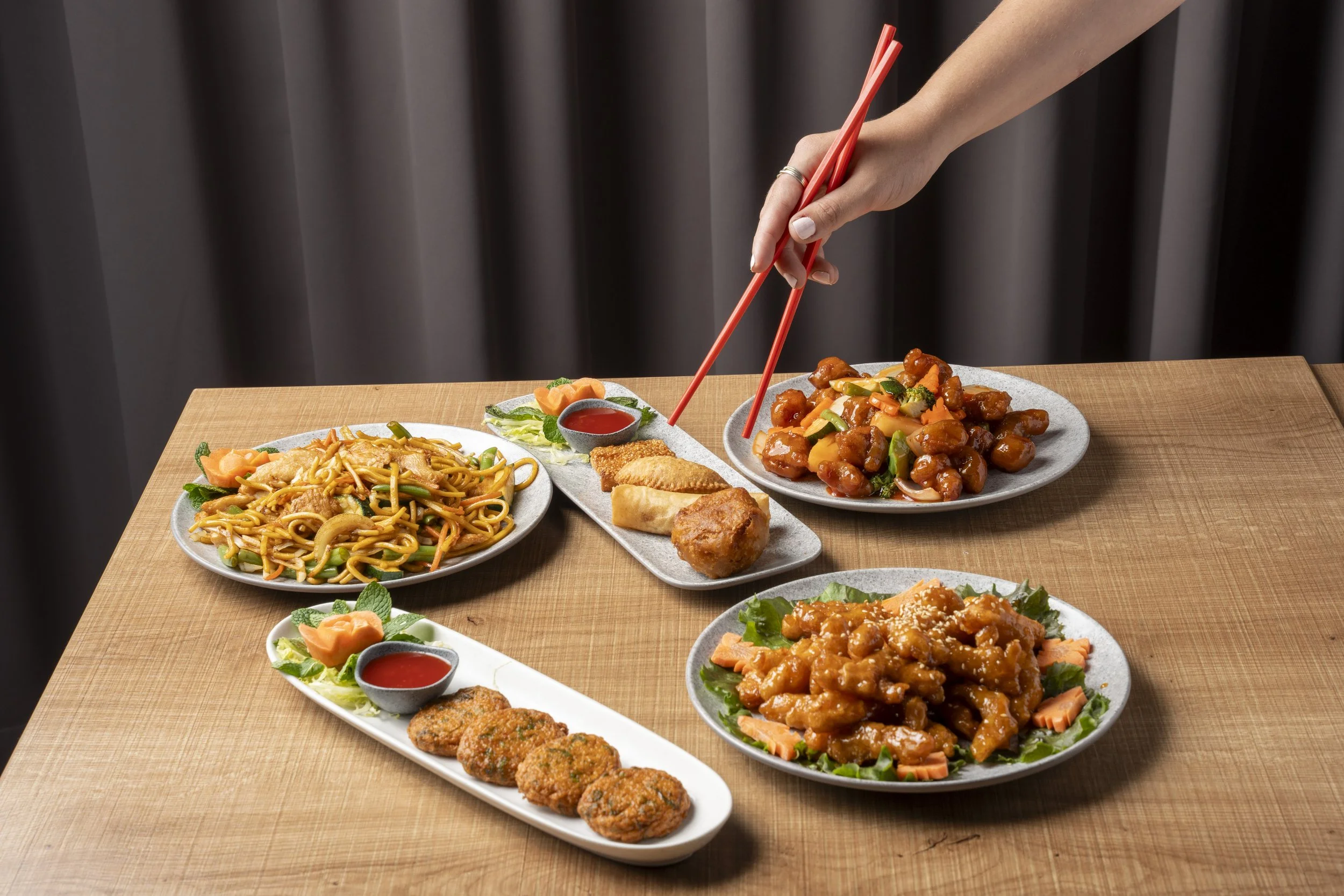 A variety of Asian dishes on a wooden table, including noodles, fried chicken, stir-fried vegetables, and sweet and sour chicken, with a person's hand using red chopsticks to pick up food. Gold Coast Food Photography by Joey Bailey