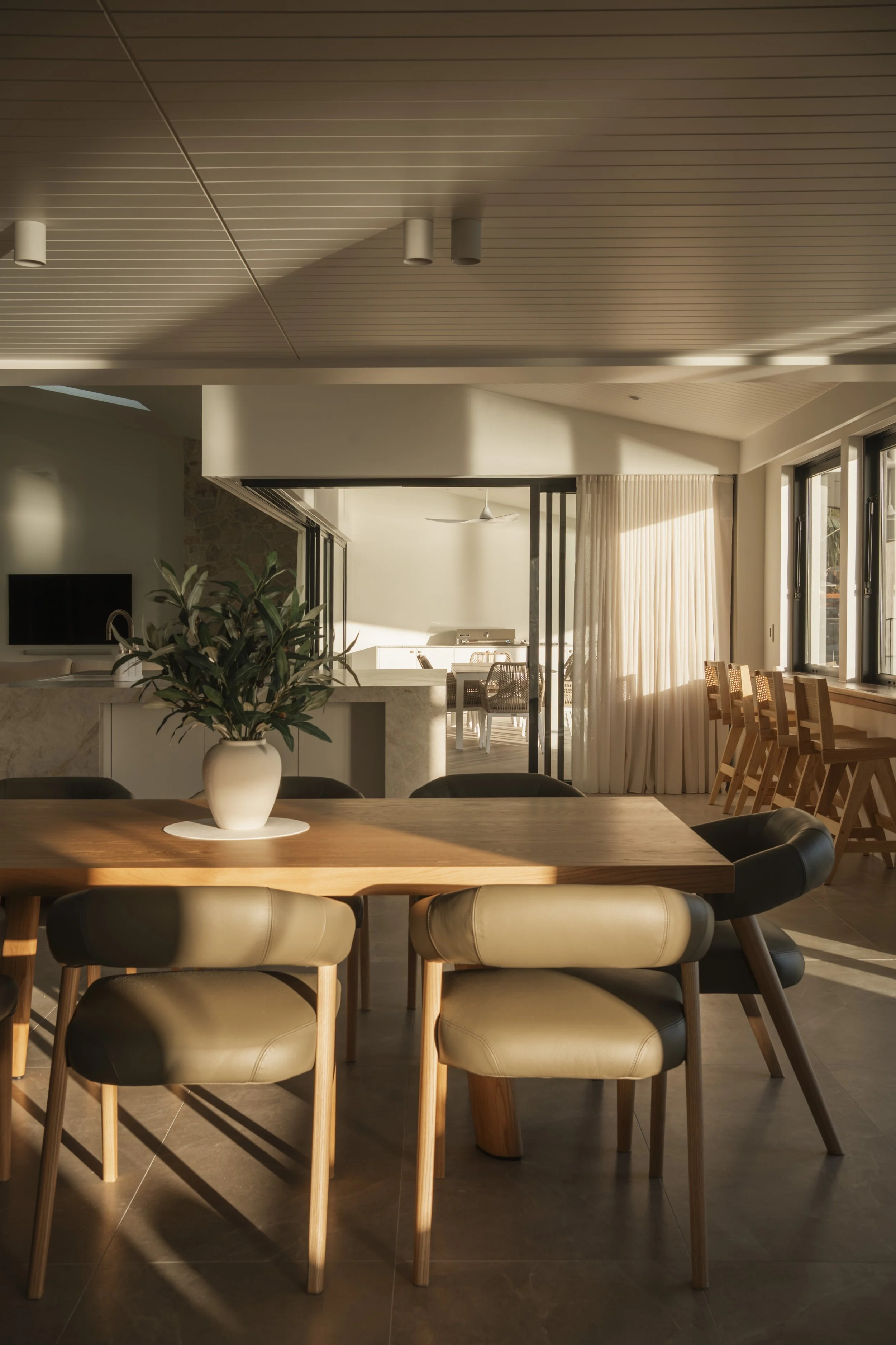 Sunlight illuminates a modern dining room with a wooden table, beige leather chairs, and a vase with green leaves. Gold Coast Architecture photography Joey Bailey
