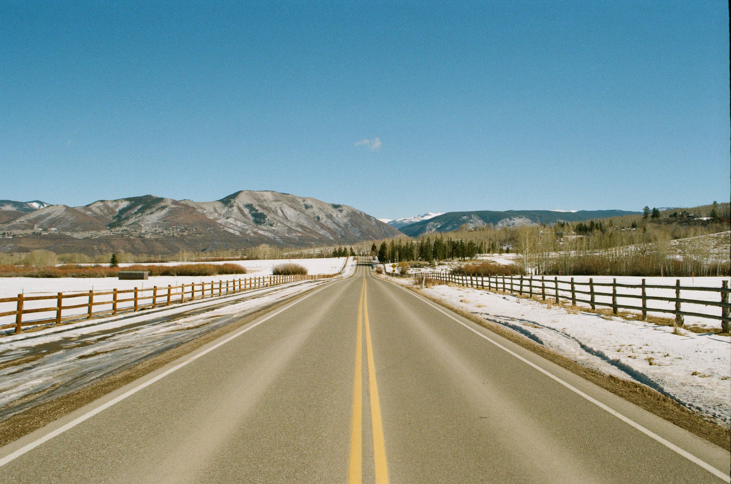A two-lane road stretches into the distance, flanked by wooden guardrails and snowy fields, with mountains in the background under a clear blue sky. Byron Bay Photography Joey Bailey