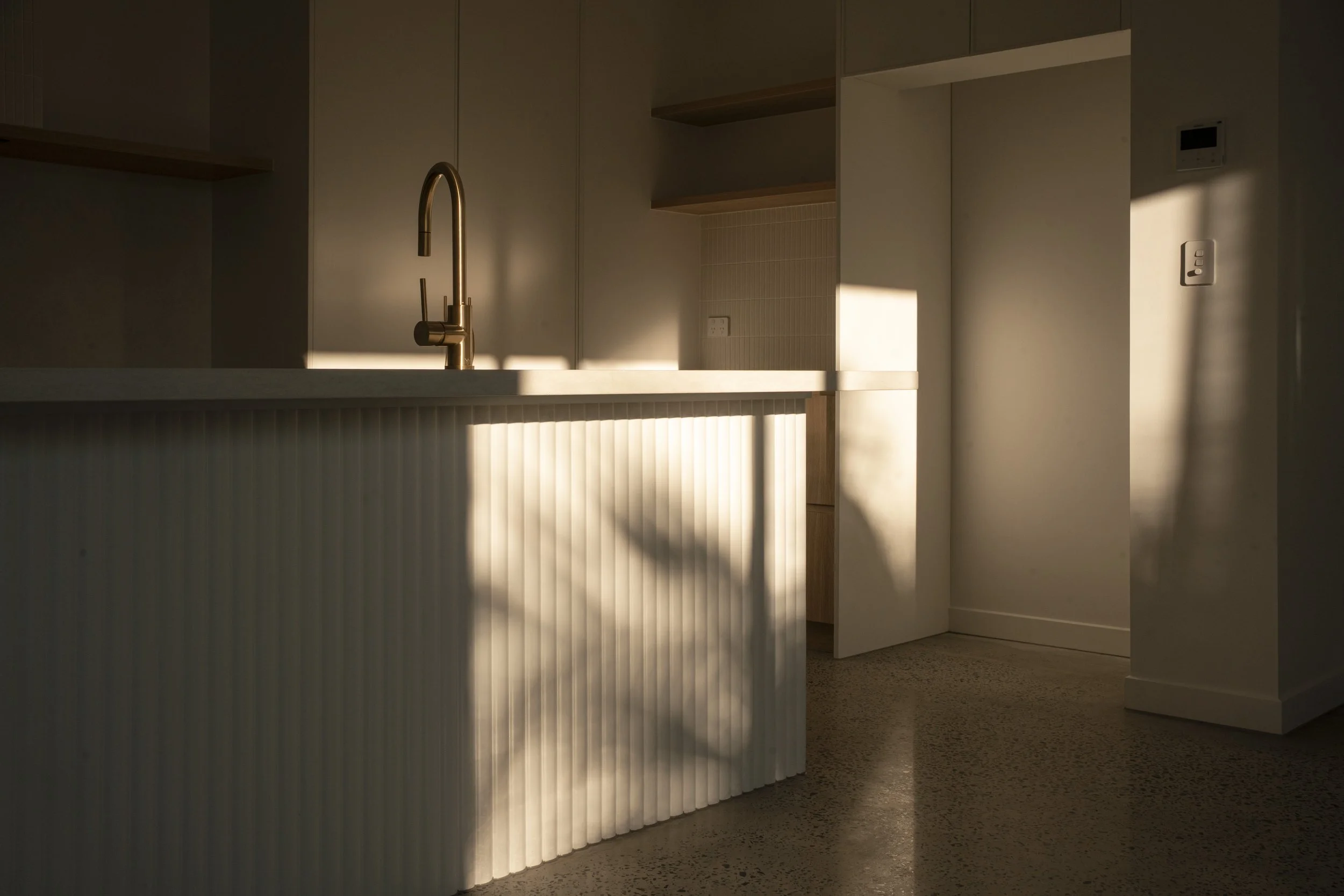 Minimalist kitchen with a gold faucet, off-white cabinetry, and sunlight casting shadows on the counter and wall. Architectural photography by Joey Bailey, Gold Coast