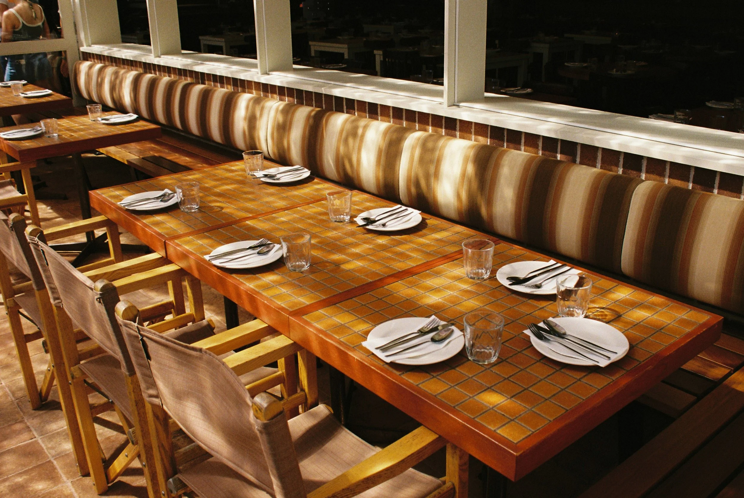 A long wooden dining table set for a meal, with striped upholstered bench seats along one side and wooden chairs on the other, holding white plates, silverware, and drinking glasses.  Club Rose Bay Photography