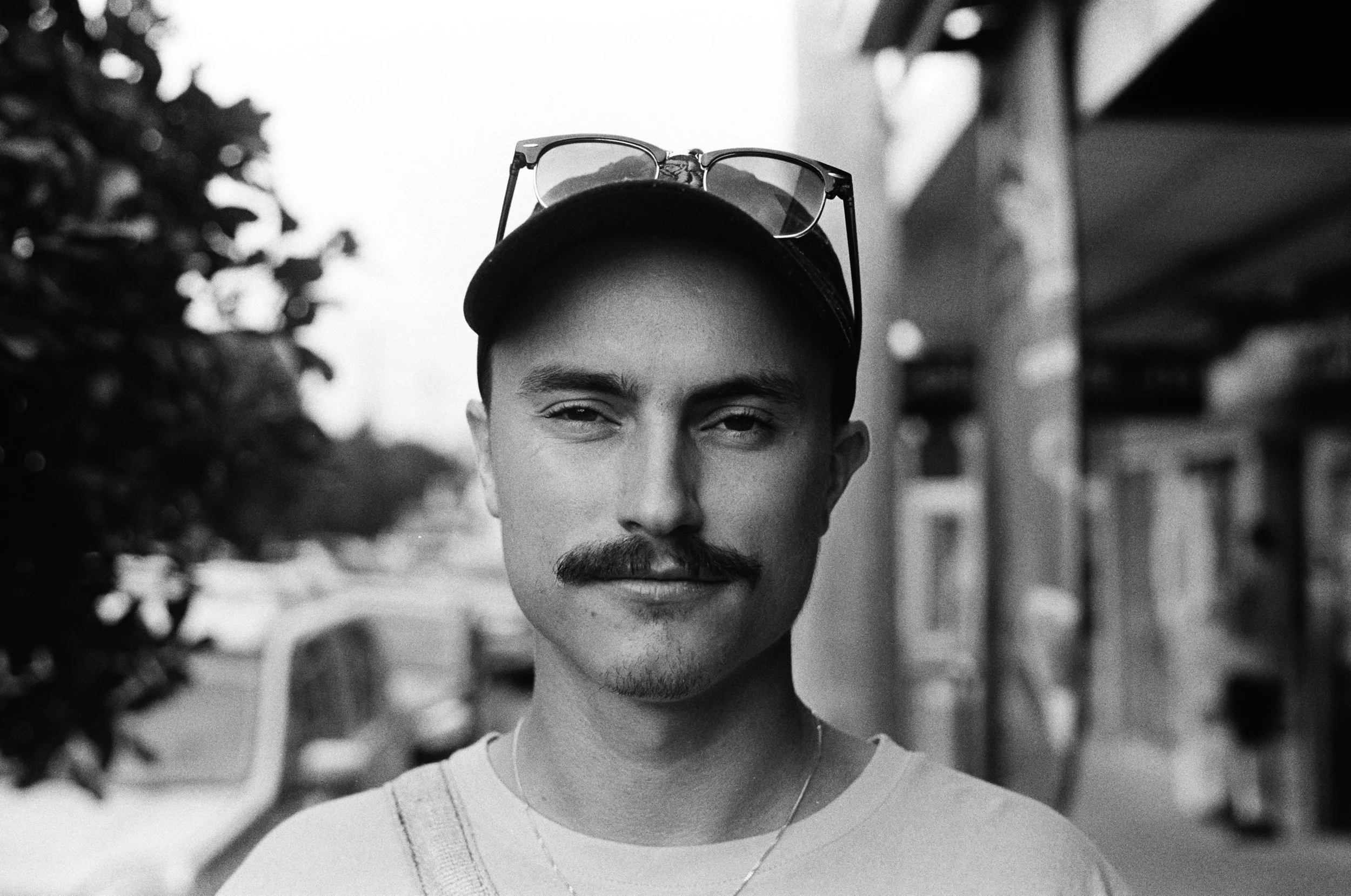 Black and white photo of a person wearing a cap with glasses on top, looking at the camera on a street.