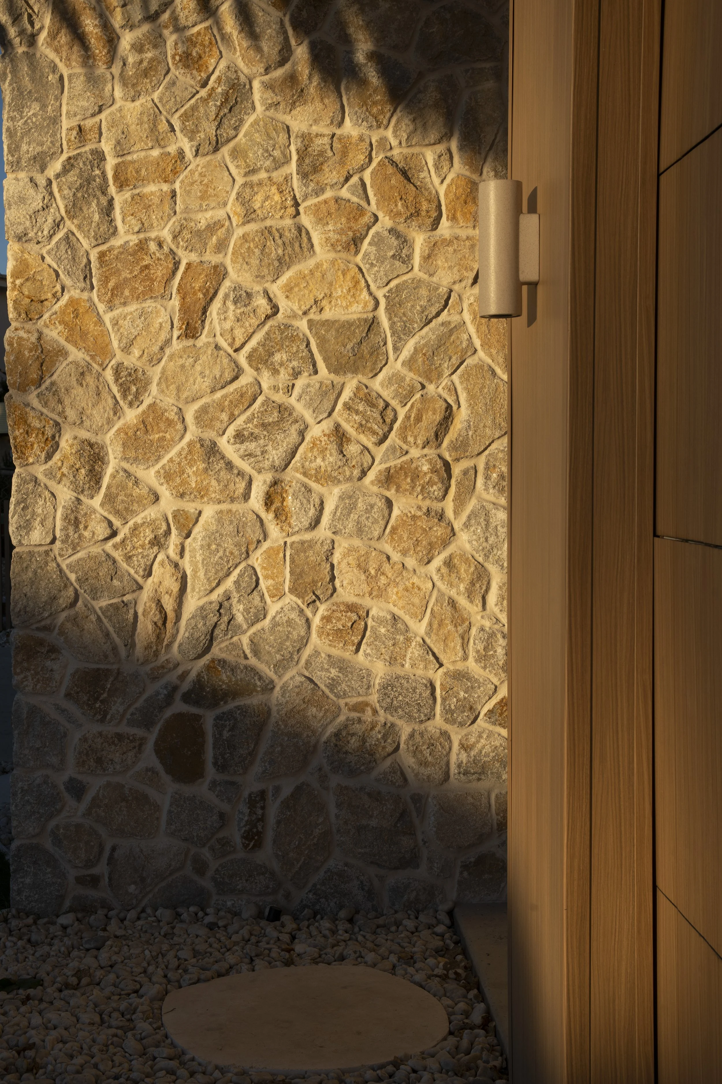 Close-up of a stone wall illuminated by sunlight next to a wooden wall with a cylindrical light fixture. Architectural photography by Joey Bailey, Gold Coast