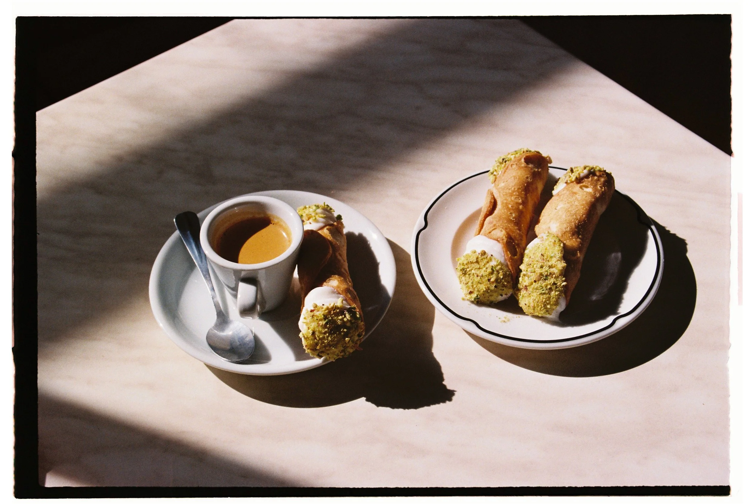 Two plates with pistachio-filled cannoli, one with a small cup of espresso and a spoon, on a marble surface with sunlight casting shadows. Byron Bay restaurant photography