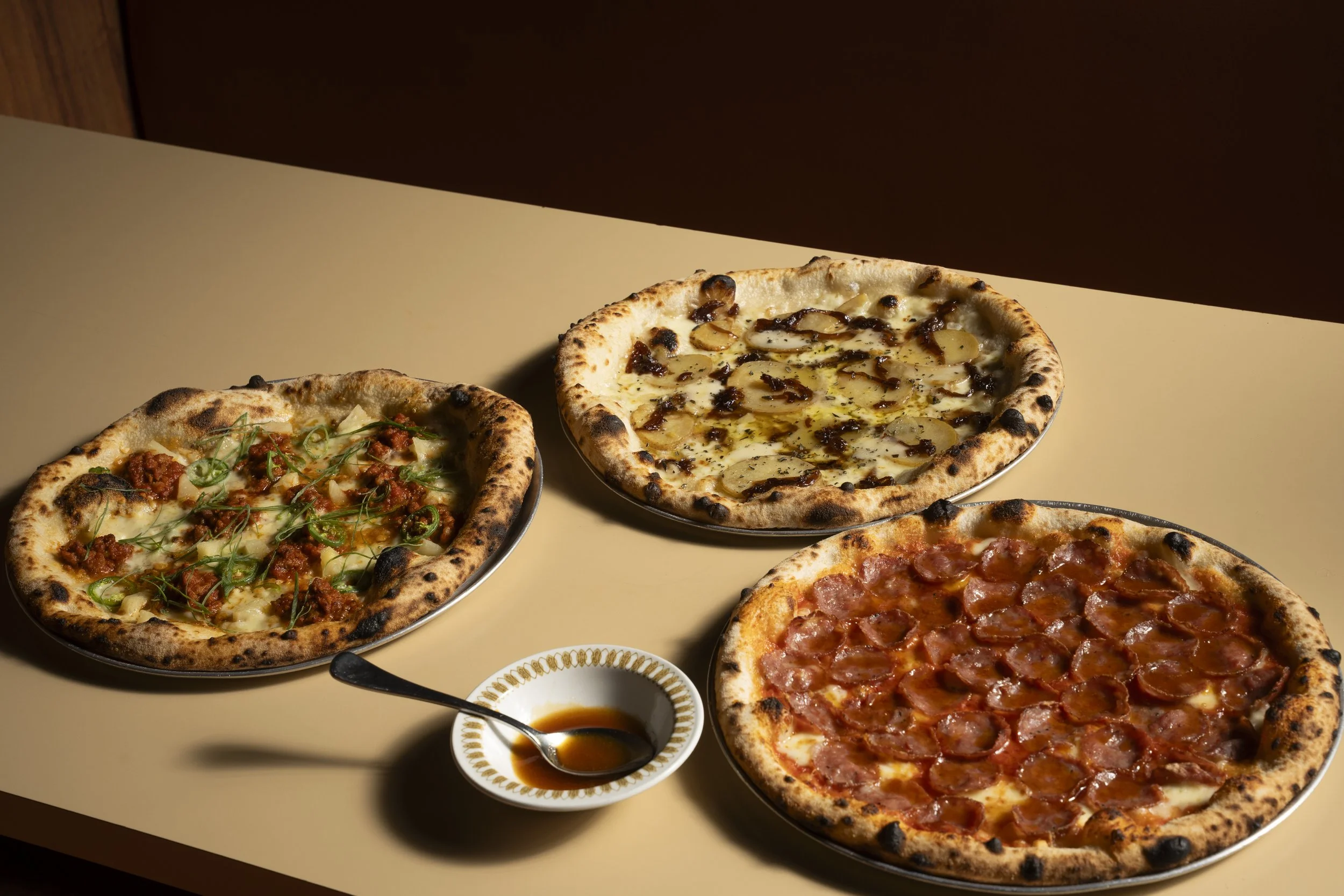 Three pizzas on a table with a bowl of dipping sauce. The pizzas include a barbecue chicken pizza, a white pizza with mushrooms, and a pepperoni pizza. Bar Evelyn Coolangatta restaurant photography Gold Coast by Joey Bailey