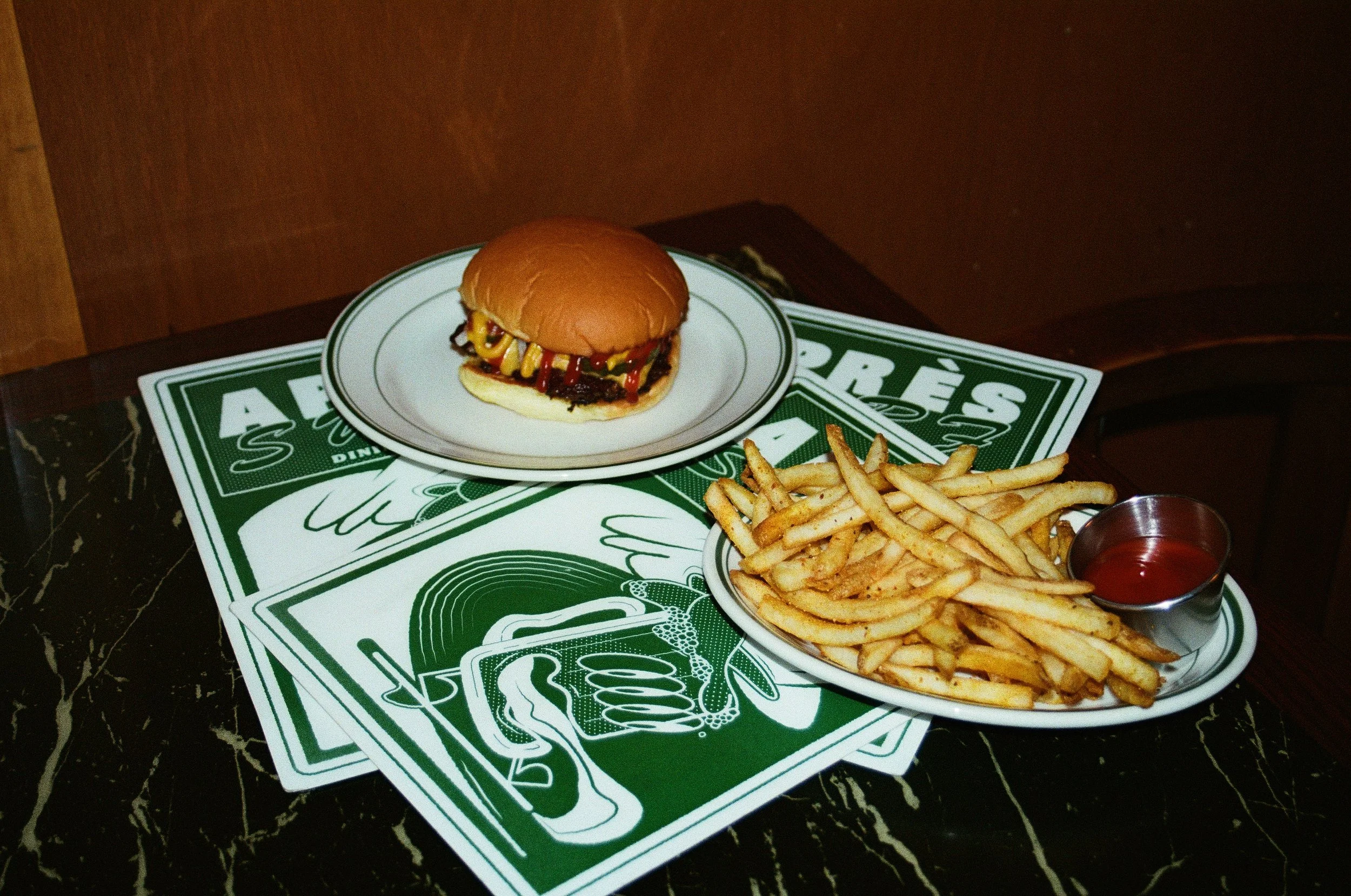 A cheeseburger with mustard and ketchup, a side of French fries, and a small cup of ketchup on a marble table, with Coca-Cola branded paper placemats underneath. Après Surf Burleigh Heads, Gold Coast – photography by Joey Bailey