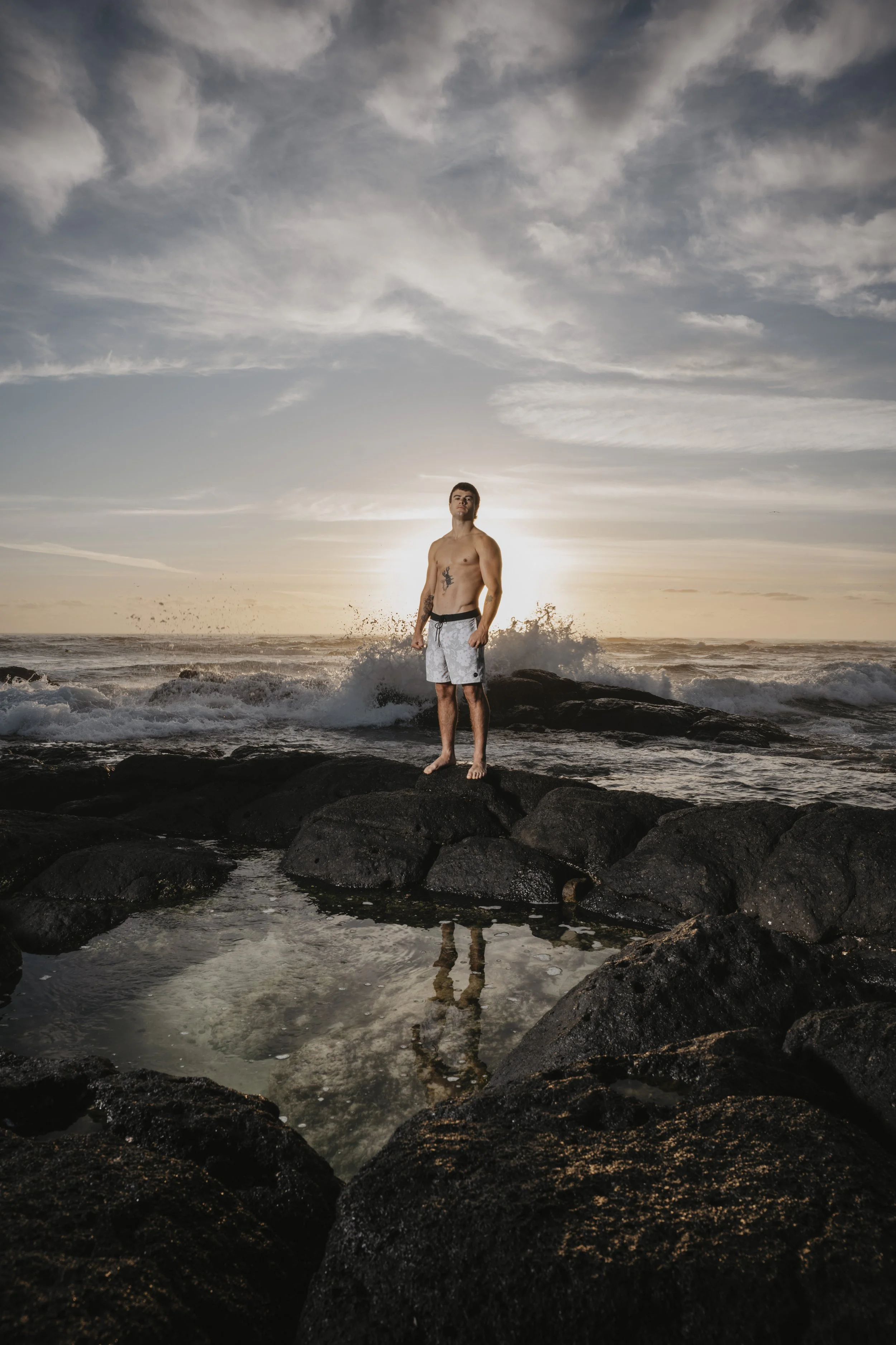 A man standing on black rocks at the beach during sunset, with waves crashing behind him and a partly cloudy sky overhead. Professional photography by Joey Bailey
