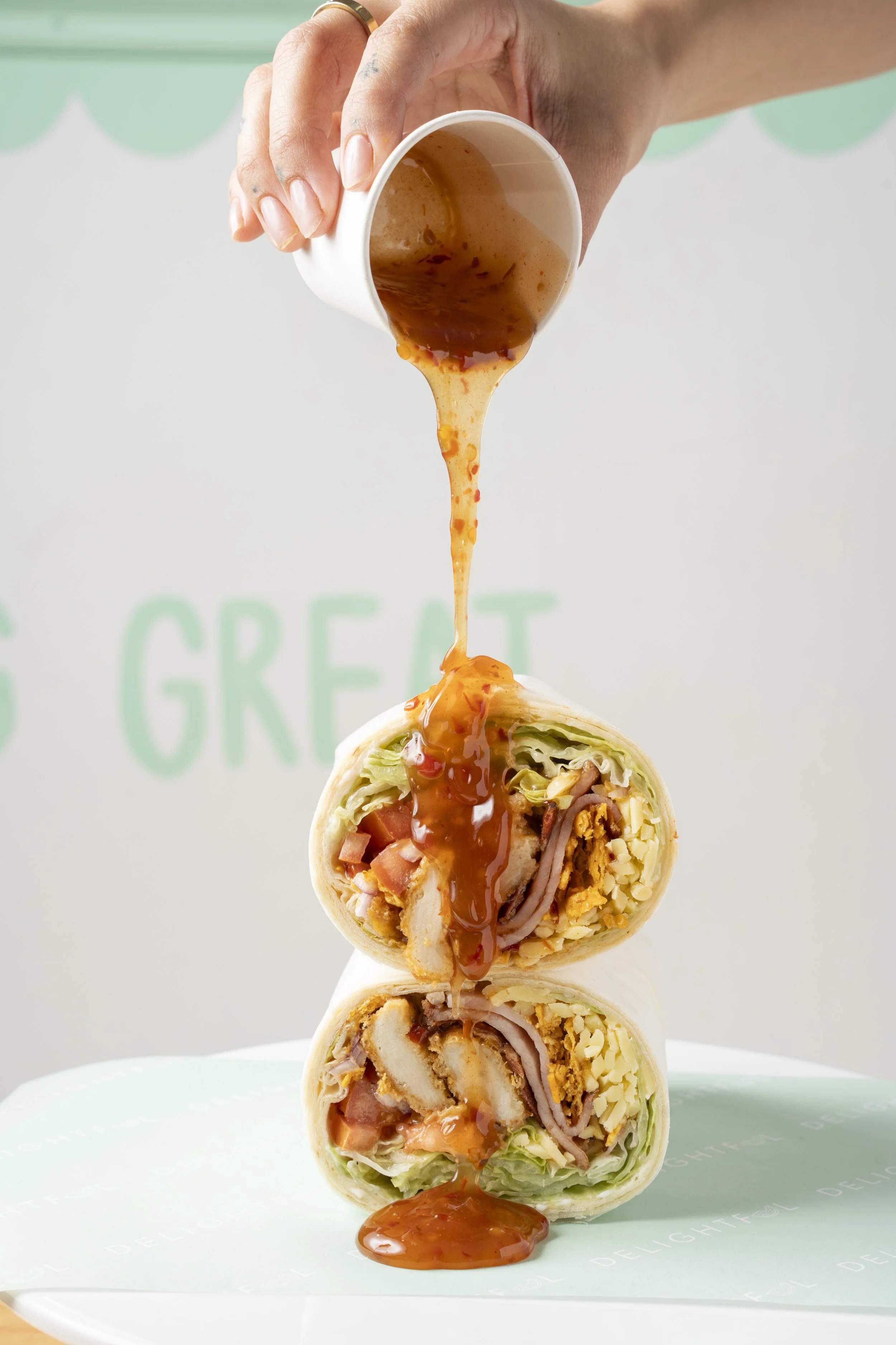 Close-up of a hand pouring sauce over a stacked chicken wrap cut in half, revealing lettuce, chicken, ham, shredded cheese, and sauce inside, on a plate with a pastel background. Gold coast food photography by Joey Bailey