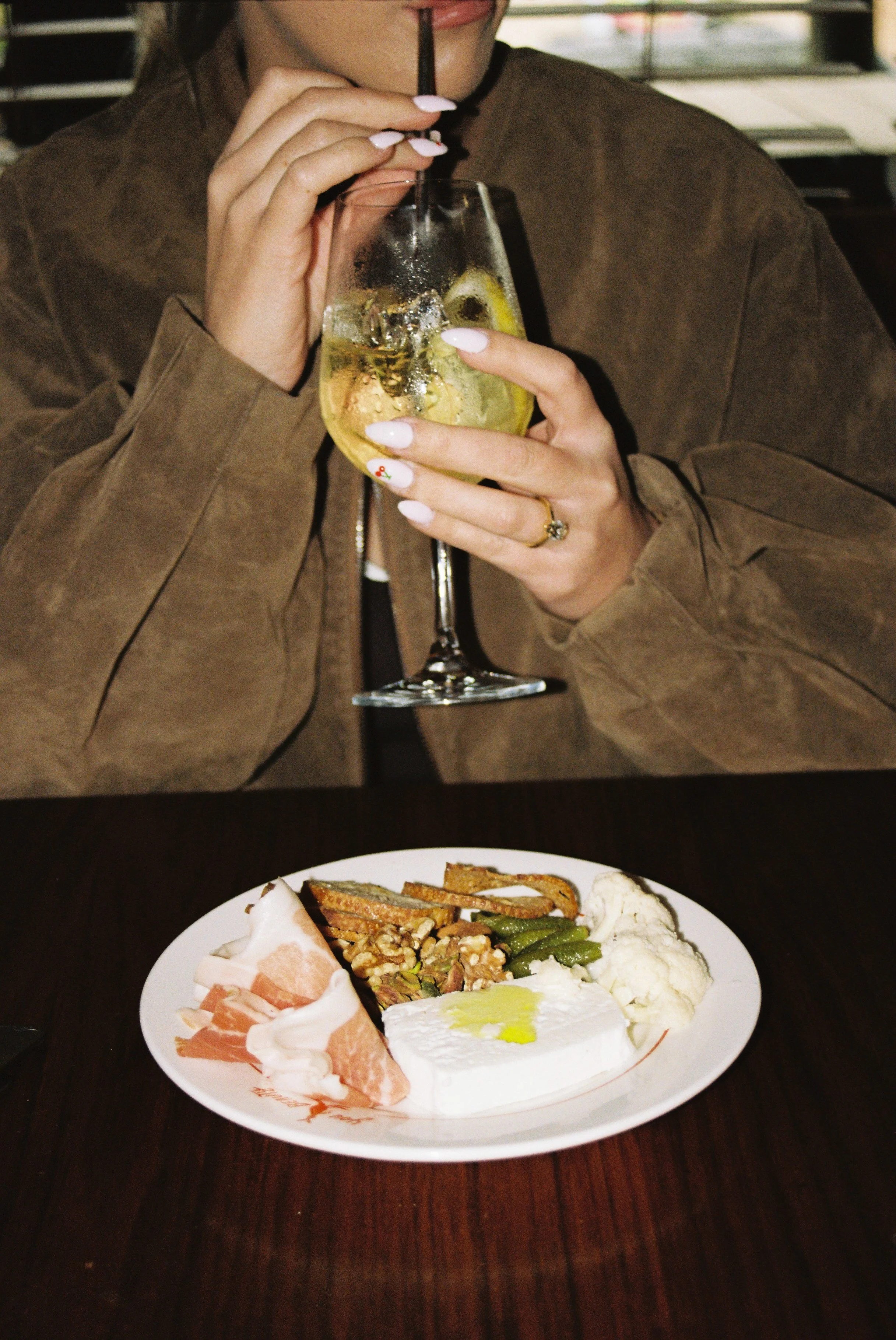 Person wearing brown jacket sipping a beverage with lemon and ice using a black straw. Plate of assorted food including sliced meats, cheese, vegetables, and dip on a dark wooden table.
