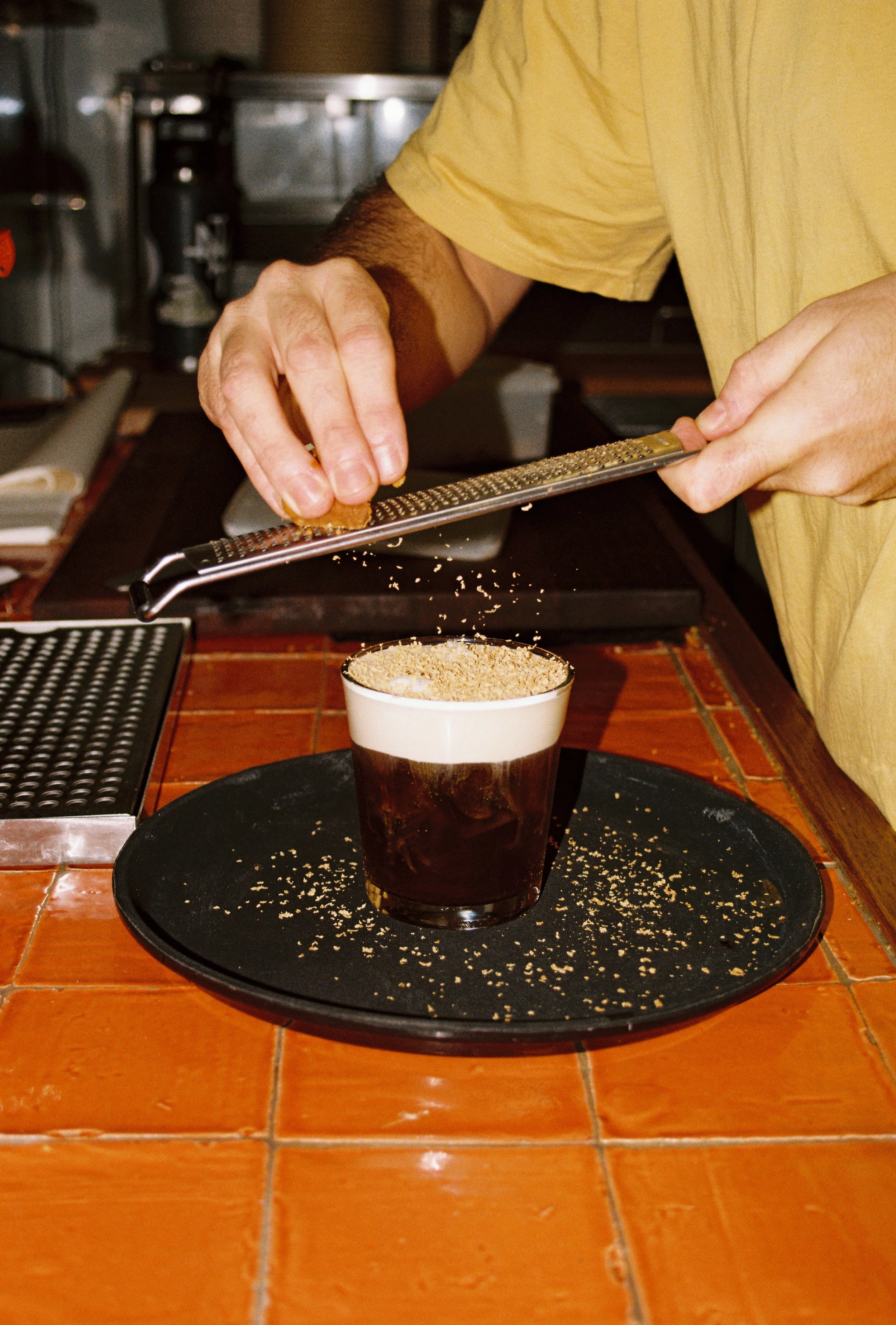 A person grating a substance over a dark beverage in a glass, placed on a black tray, on a tiled bar or countertop. Byron Bay food photography