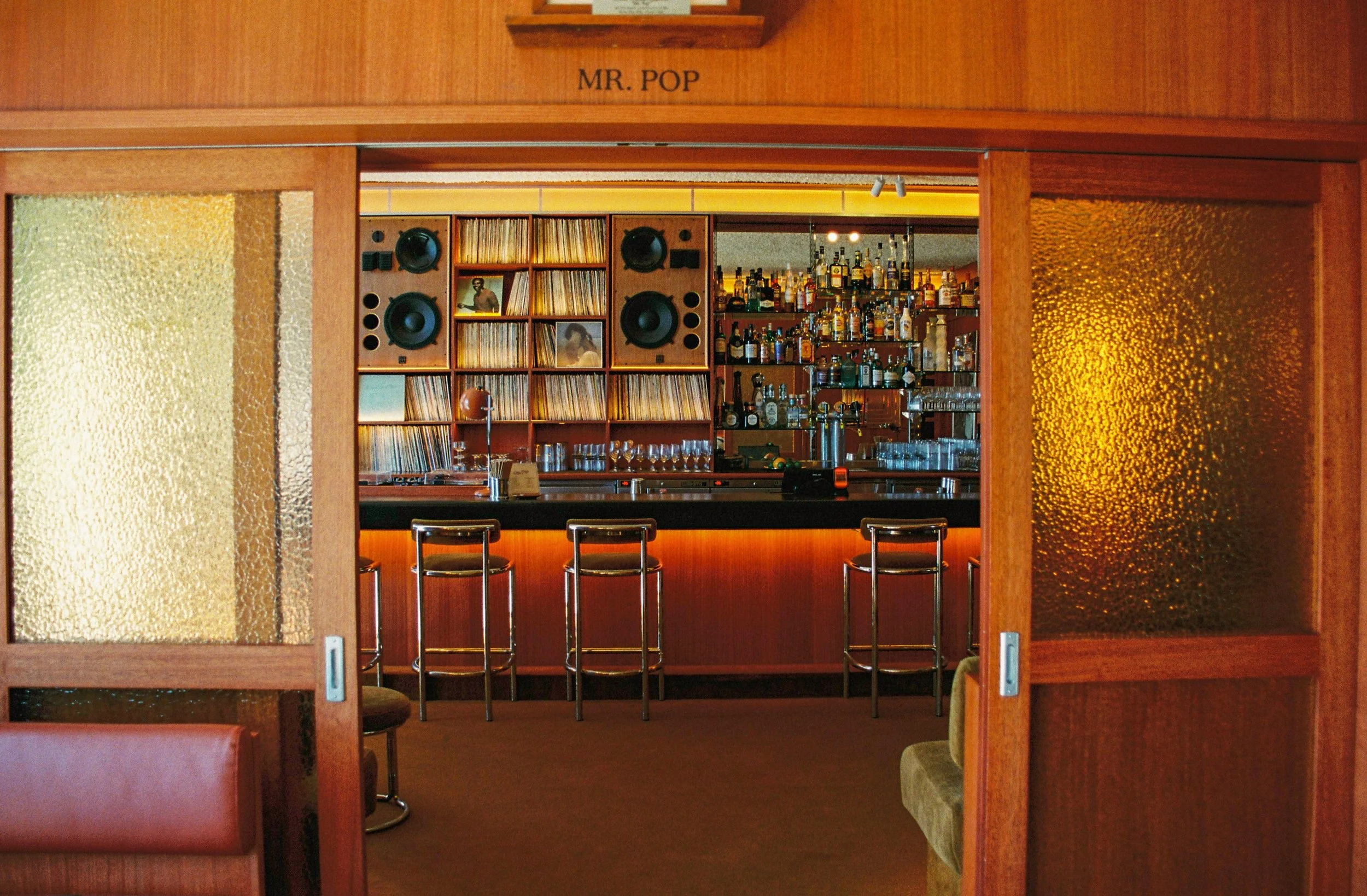 Bar area viewed through wooden sliding doors with textured glass panels, featuring a collection of liquor bottles, glasses, and bar equipment, with wood-paneled walls and framed photographs. Interior Photography Gold Coast