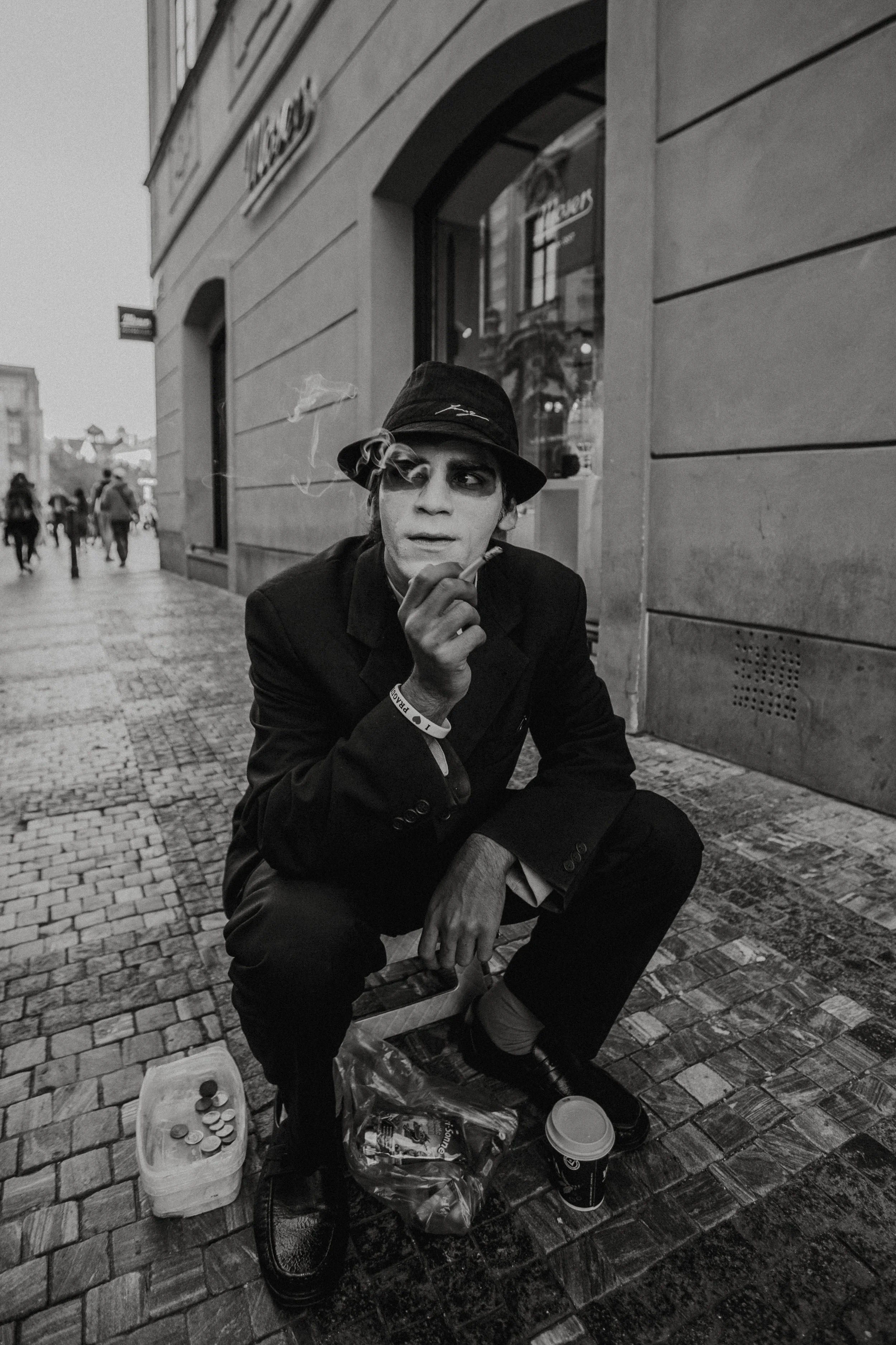 A man in a black suit and hat squatting on a cobblestone street, smoking a cigarette. He has theatrical makeup on his face. Beside him, there's a container with coins and a plastic bag with items. The scene is in black and white.