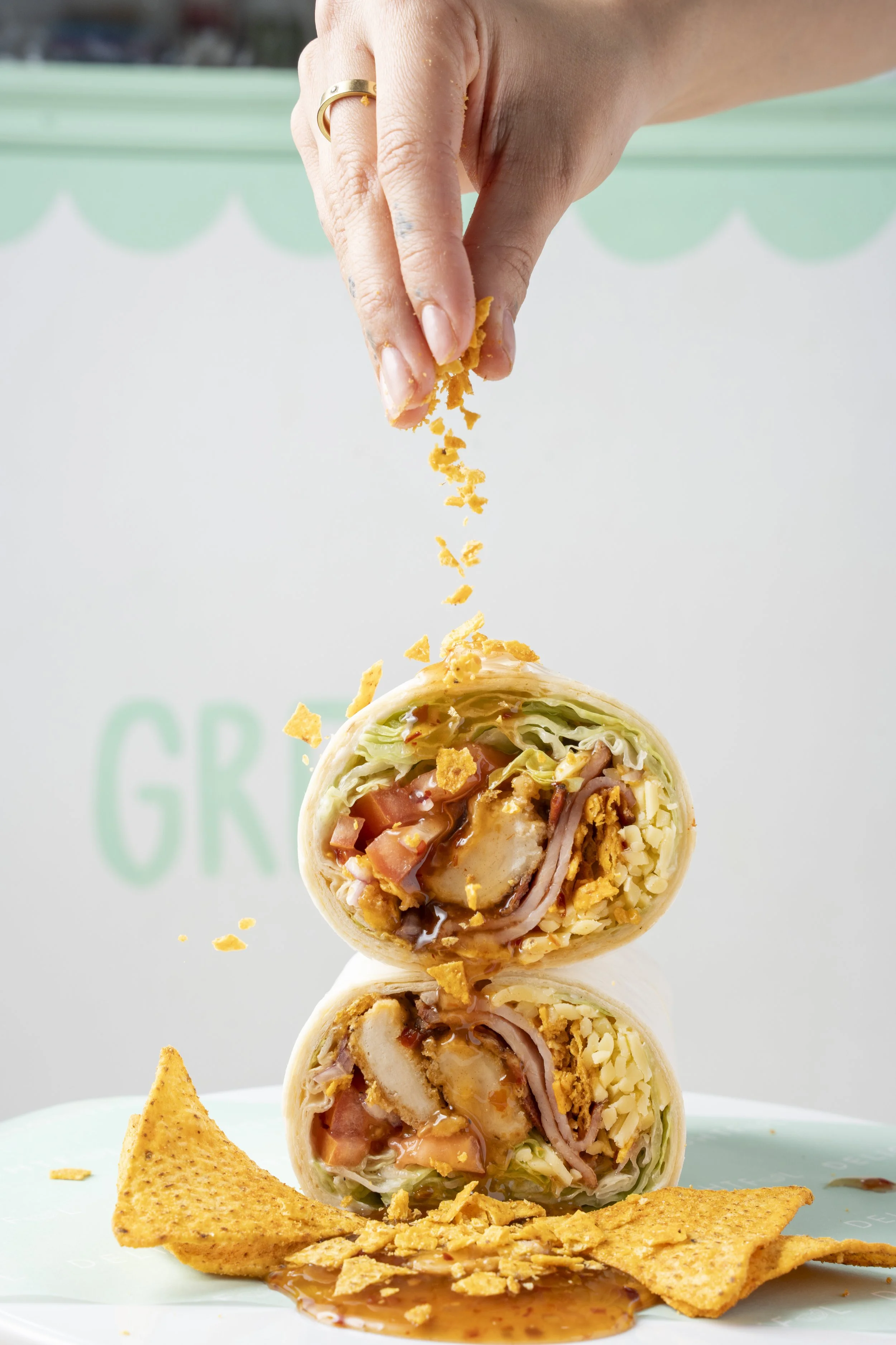 A hand sprinkling crushed chips onto a stack of cut-in-half burritos filled with lettuce, tomatoes, chicken, cheese, and sauce, with chips and sauce on a plate. Gold coast food photography by Joey Bailey