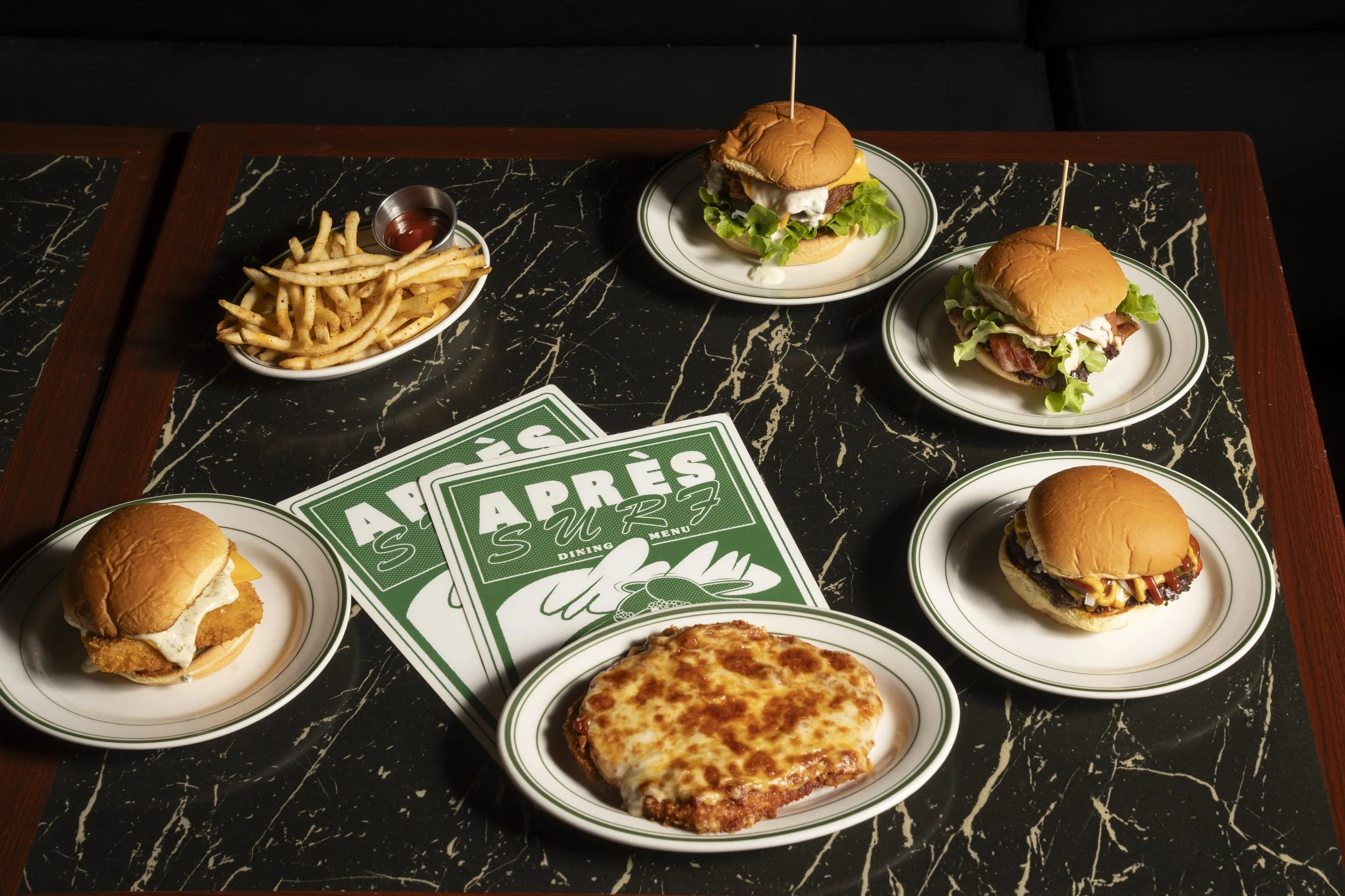 Assortment of burgers and fries on a table with two "Après Surf" menus. Each plate holds a different type of burger, and there's a dish of fries with a cup of ketchup. A cheesy baked dish is also present. Gold Coast | Byron Bay Food Photography by Jo