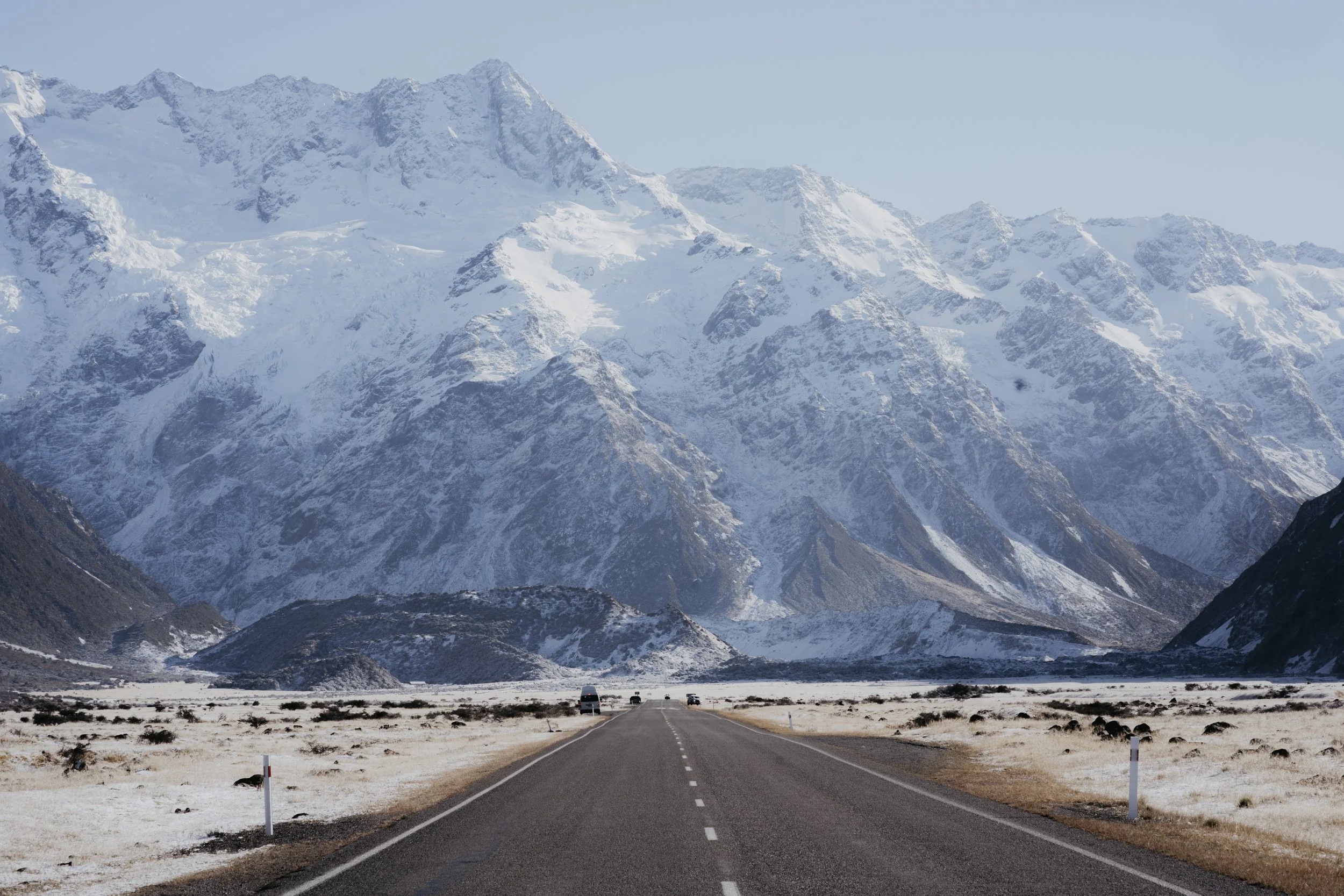 A straight two-lane road stretches through a snowy plain toward snow-covered mountains in the distance. 