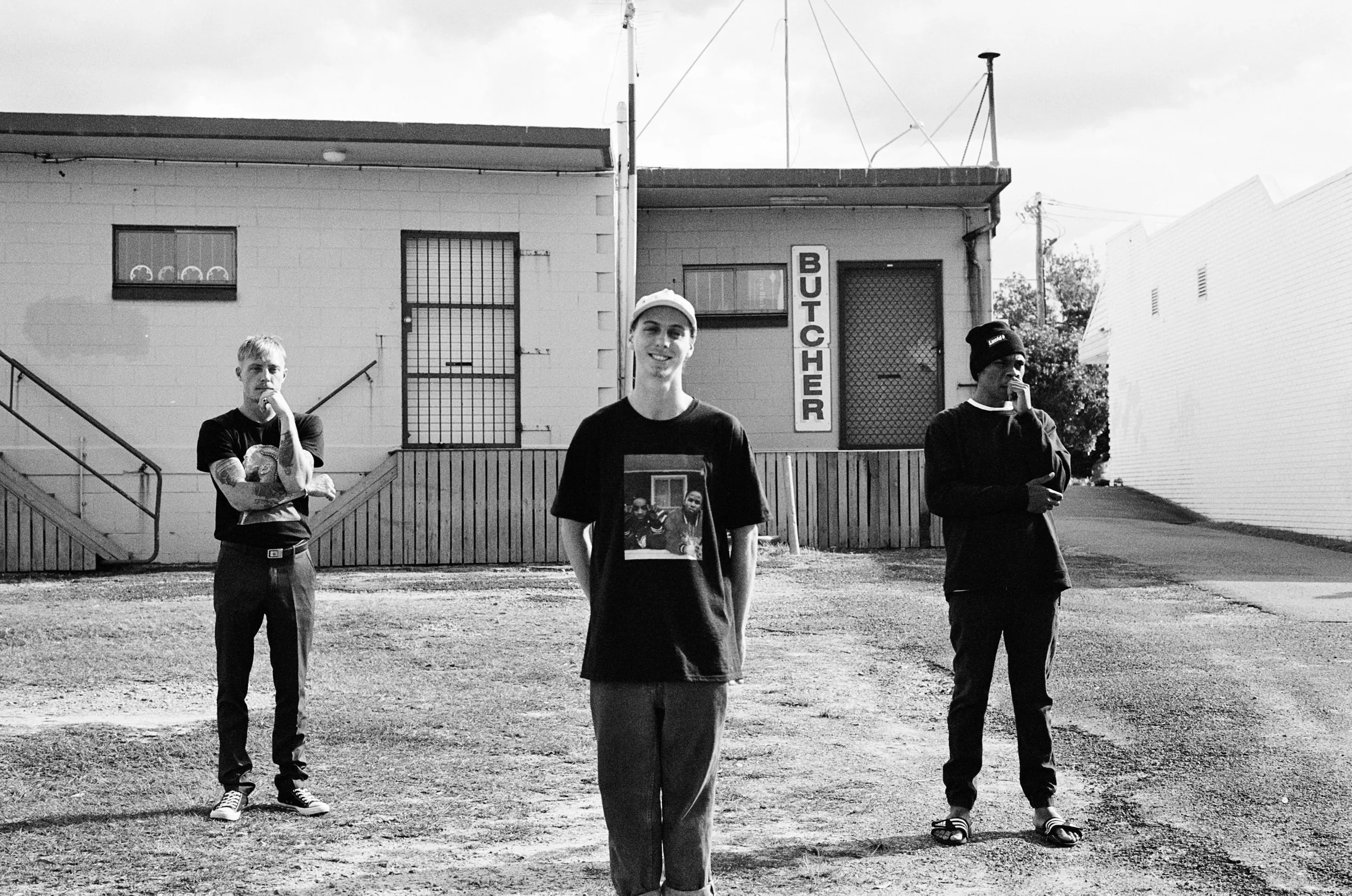 Three men standing in front of a building with a "Butcher" sign, wearing casual clothing. The backdrop includes a cinder block wall with barred windows.