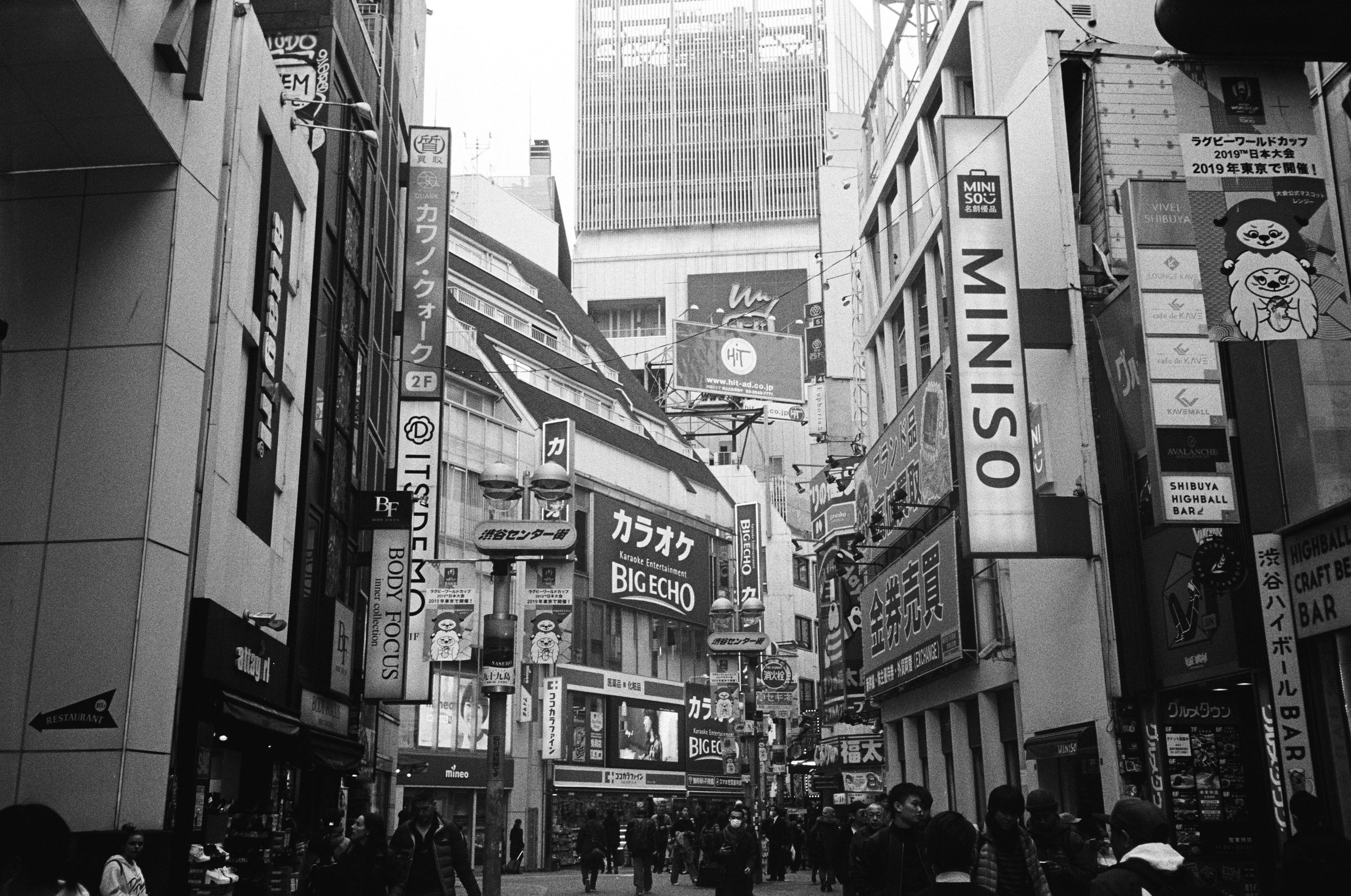 A monochrome street scene in a busy urban area, lined with commercial buildings and various signage in both Japanese and English, including brands like Miniso. People are walking along the street, and advertising banners are visible.