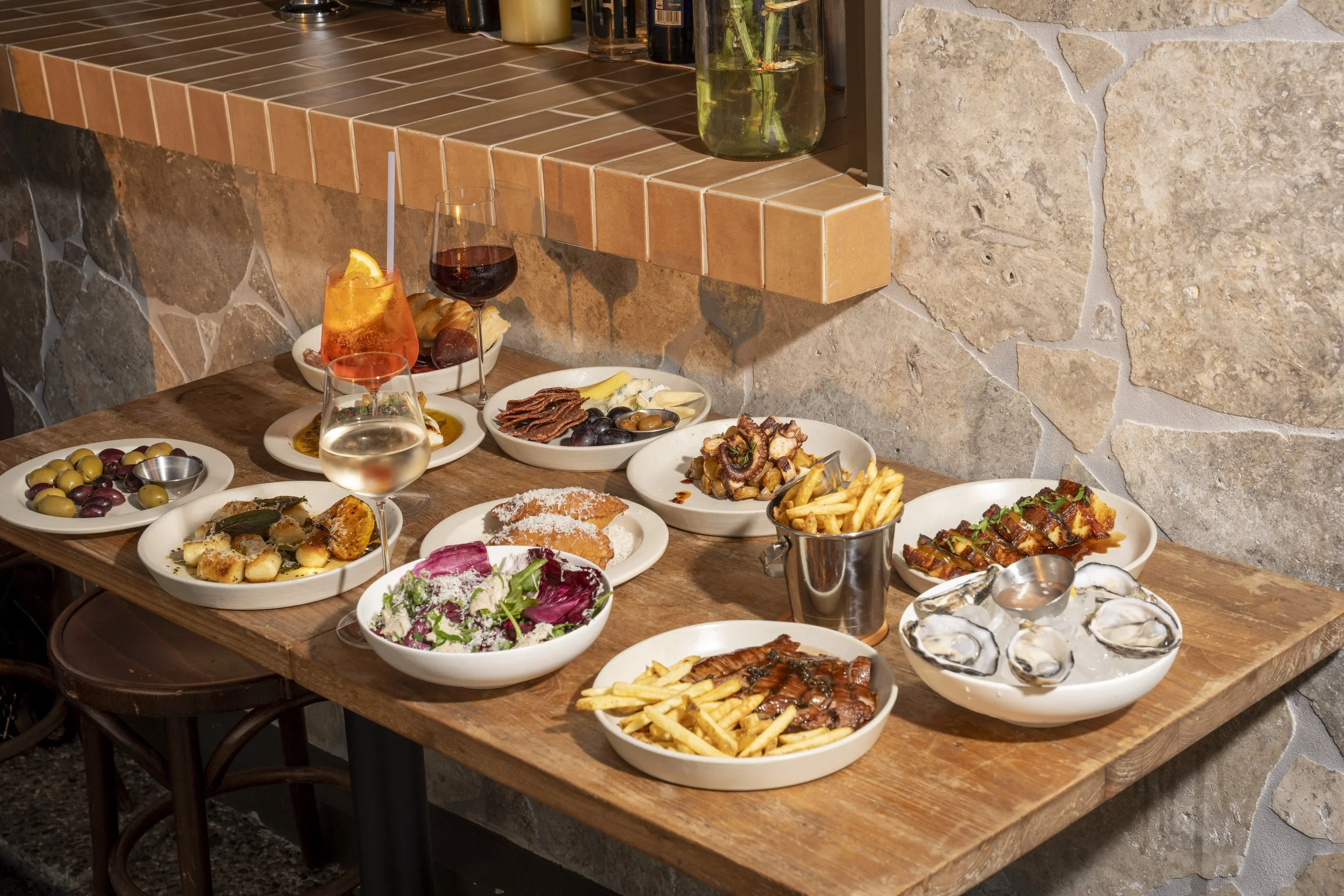 A wooden table set with a variety of dishes including salads, olives, oysters, fries, grilled meat, and desserts, with wine and other beverages. Gold Coast Food Photography by Joey Bailey