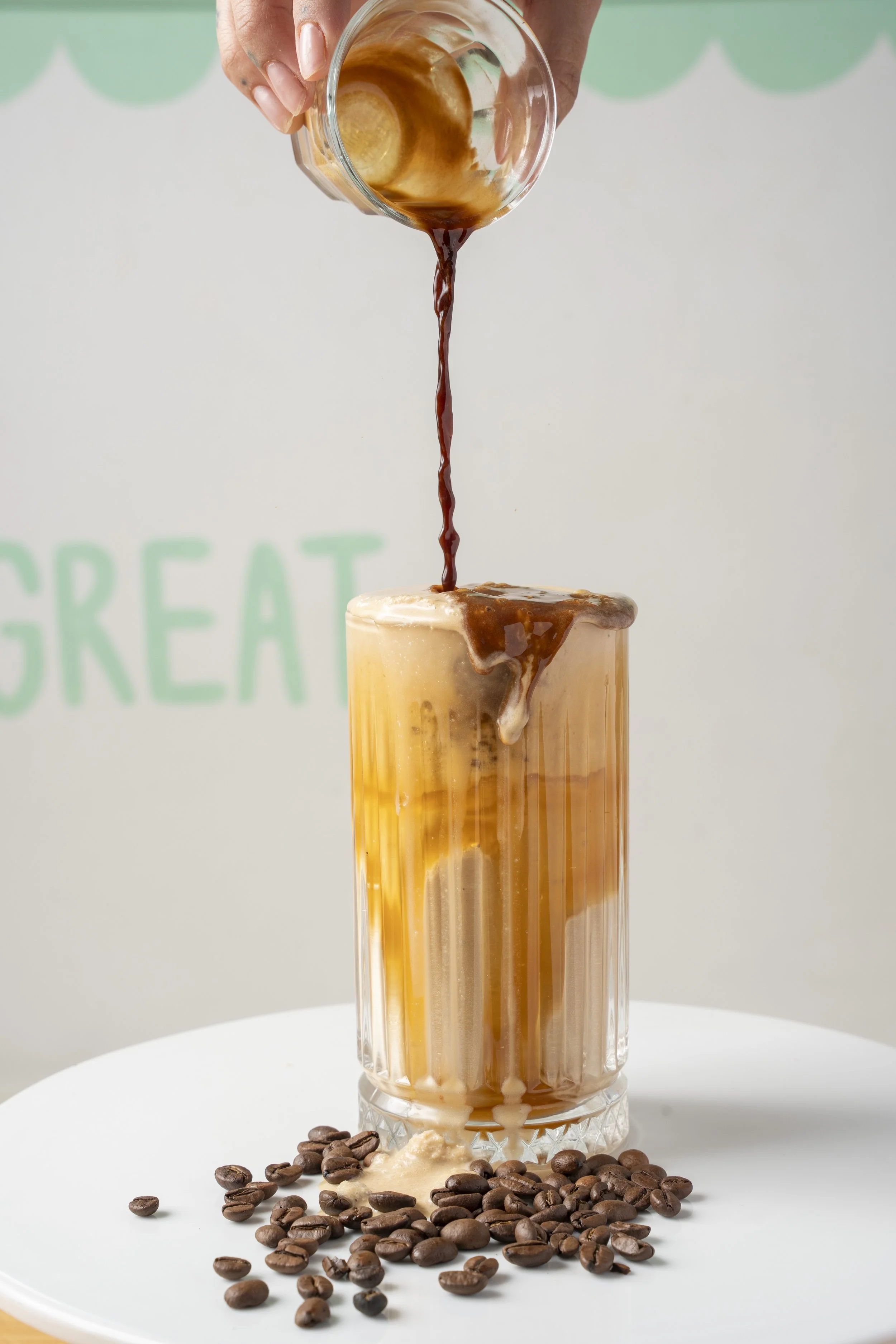 A hand pouring coffee into a tall glass of iced coffee, surrounded by scattered coffee beans on a white surface. Gold Coast Food photography by Joey Bailey