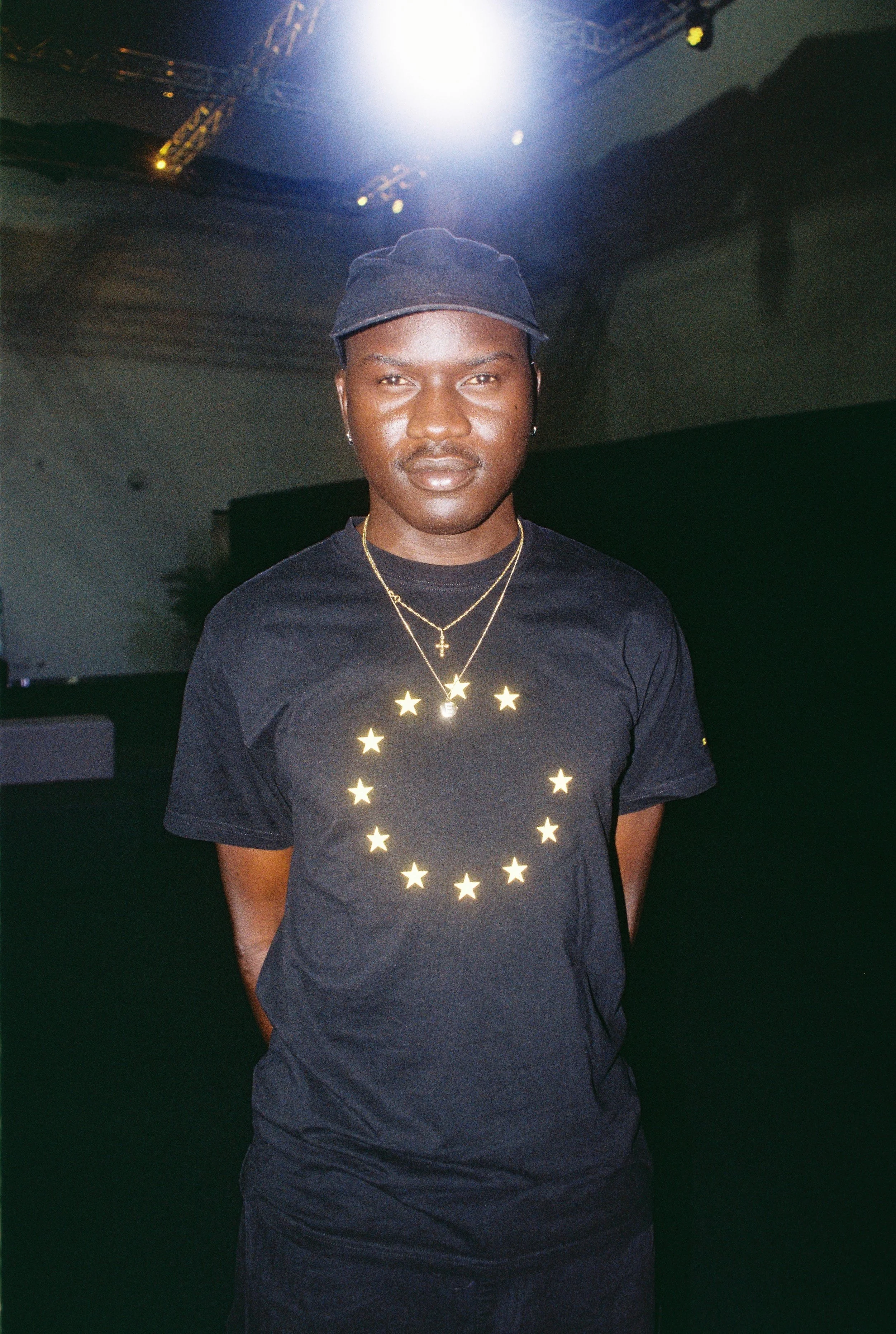 Skin on Skin wearing a black t-shirt with circular star pattern and a cap, standing under bright overhead light.