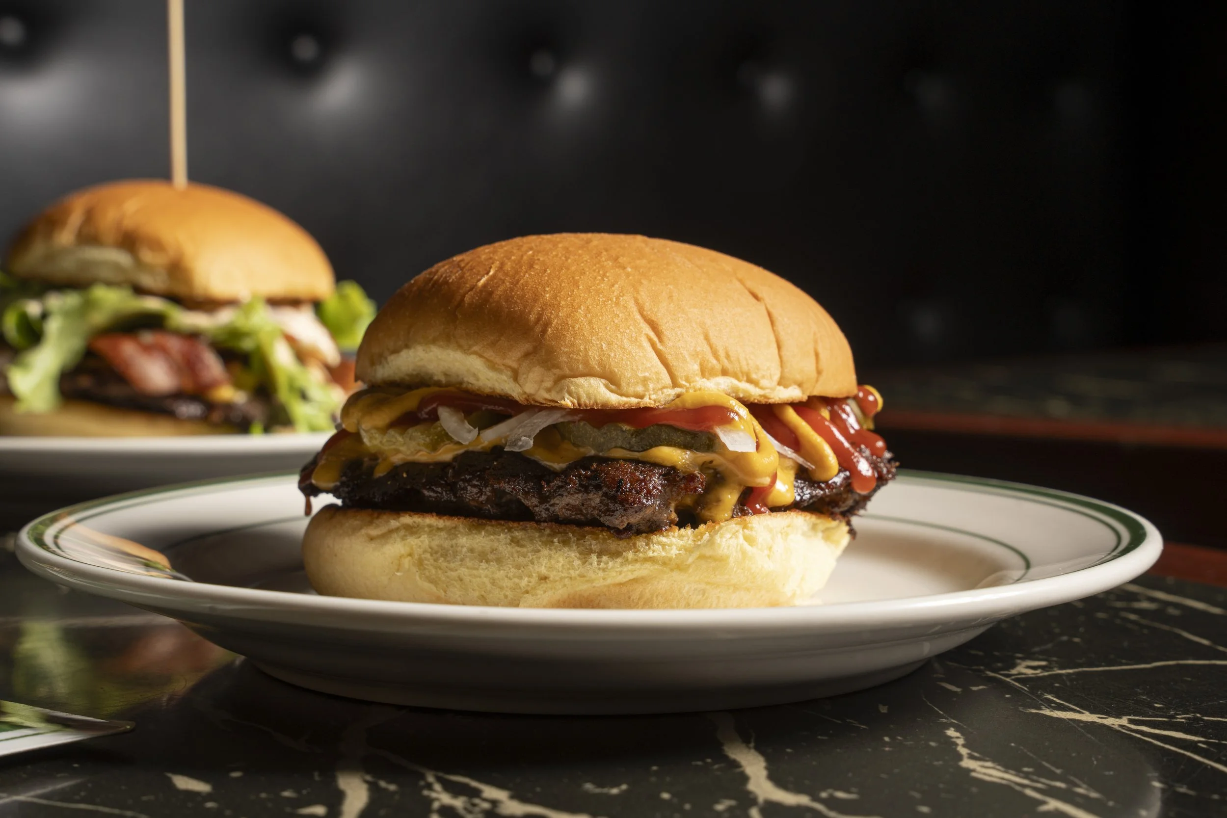 Close-up of a cheeseburger with pickles, onions, ketchup, and mustard on a white plate; another burger is blurred in the background on a marble table.Gold Coast | Byron Bay Food Photography by Joey Bailey