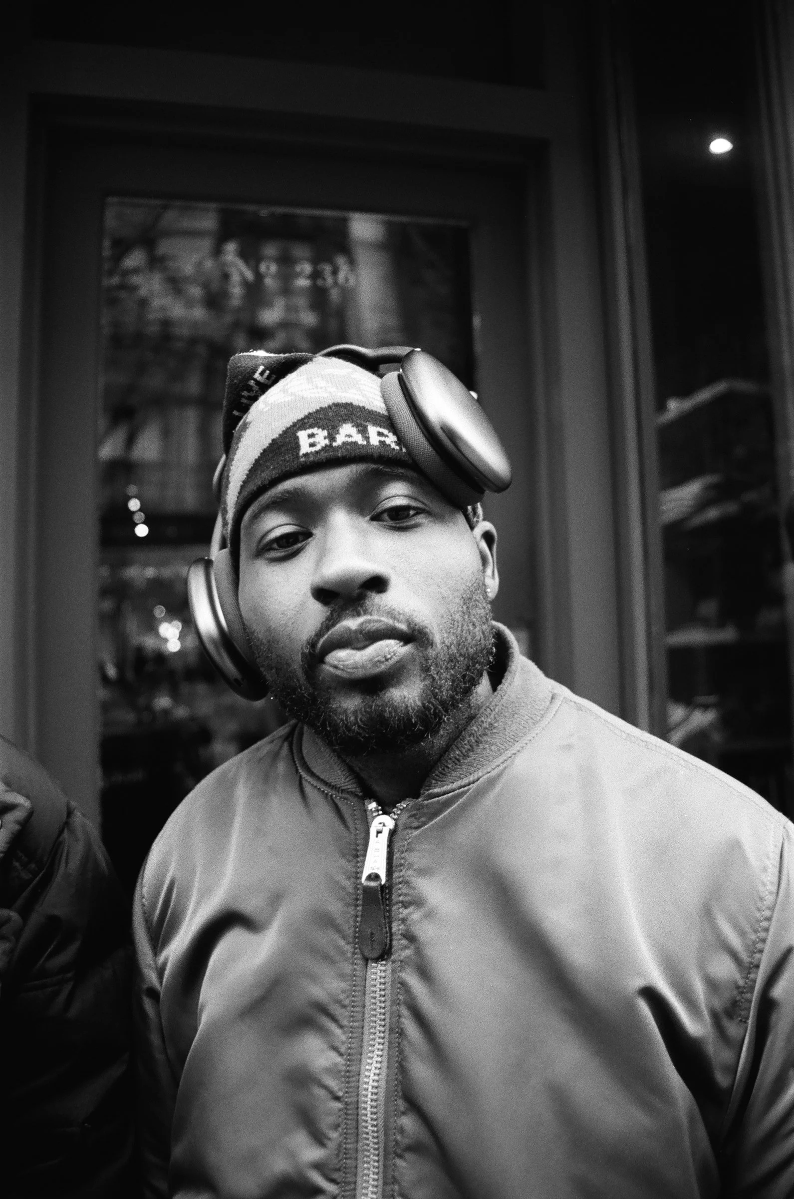 Black and white photo of a man wearing headphones around his neck and a beanie, standing in front of a glass door.