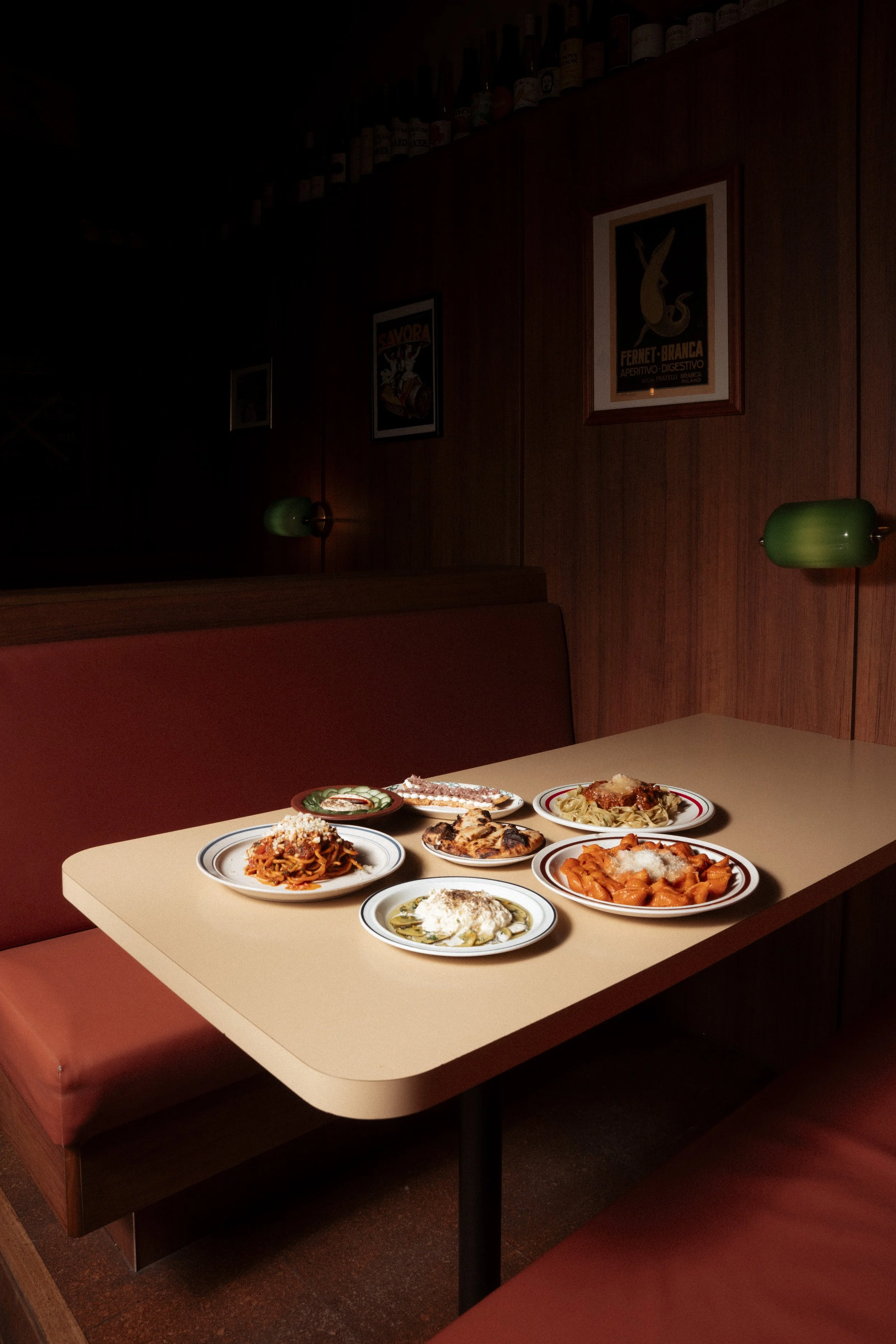 A beige table with six plates of various pasta dishes in a dimly lit restaurant with wooden wall panels and green wall-mounted lamps. Gold Coast Food photography by Joey Bailey