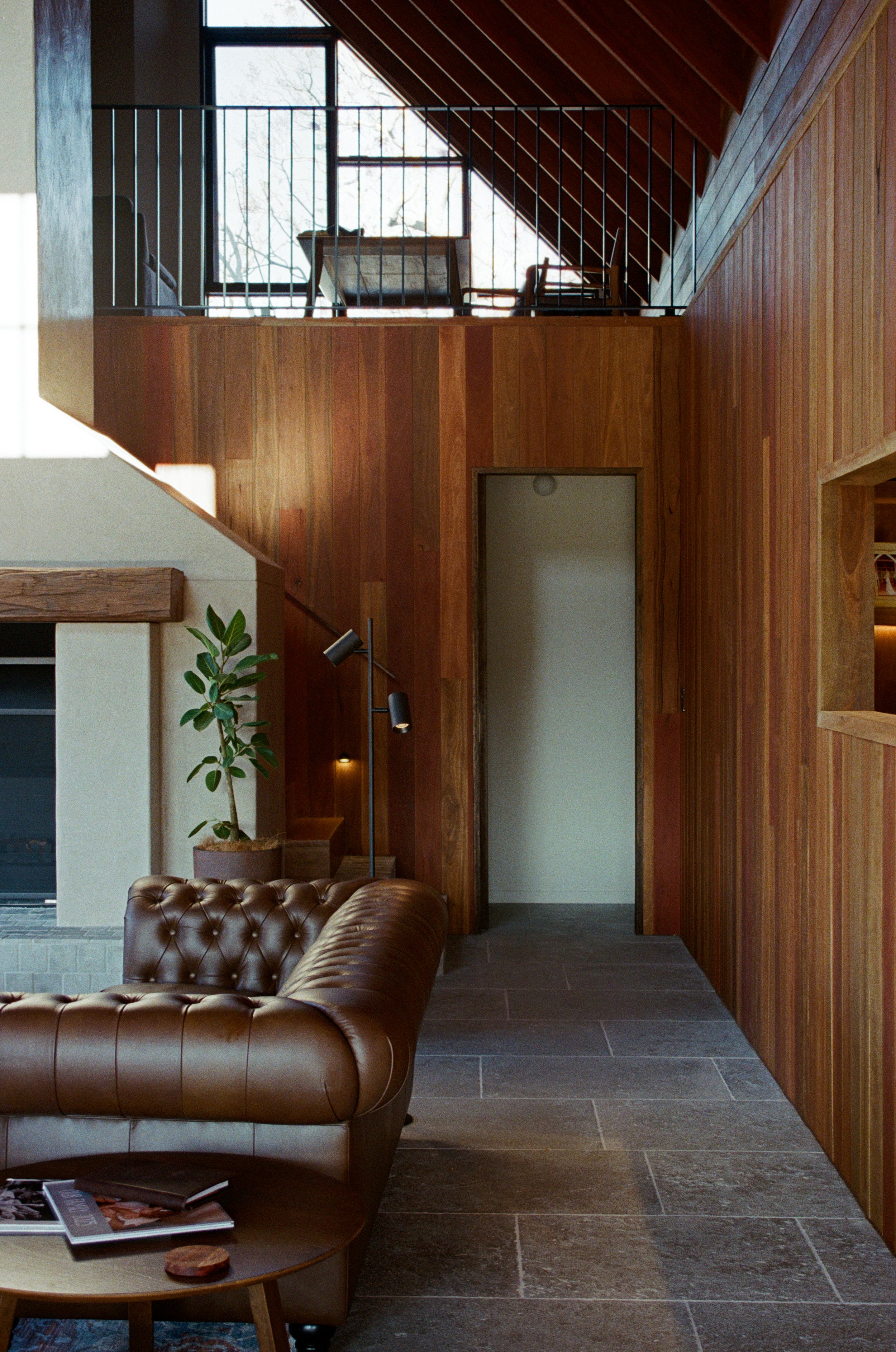 Interior view of a modern living room with wooden walls, a leather sofa, a small table with magazines, and a potted plant, with a stairway leading to a second-floor balcony, Gold Coast Interior Photography by Joey Bailey