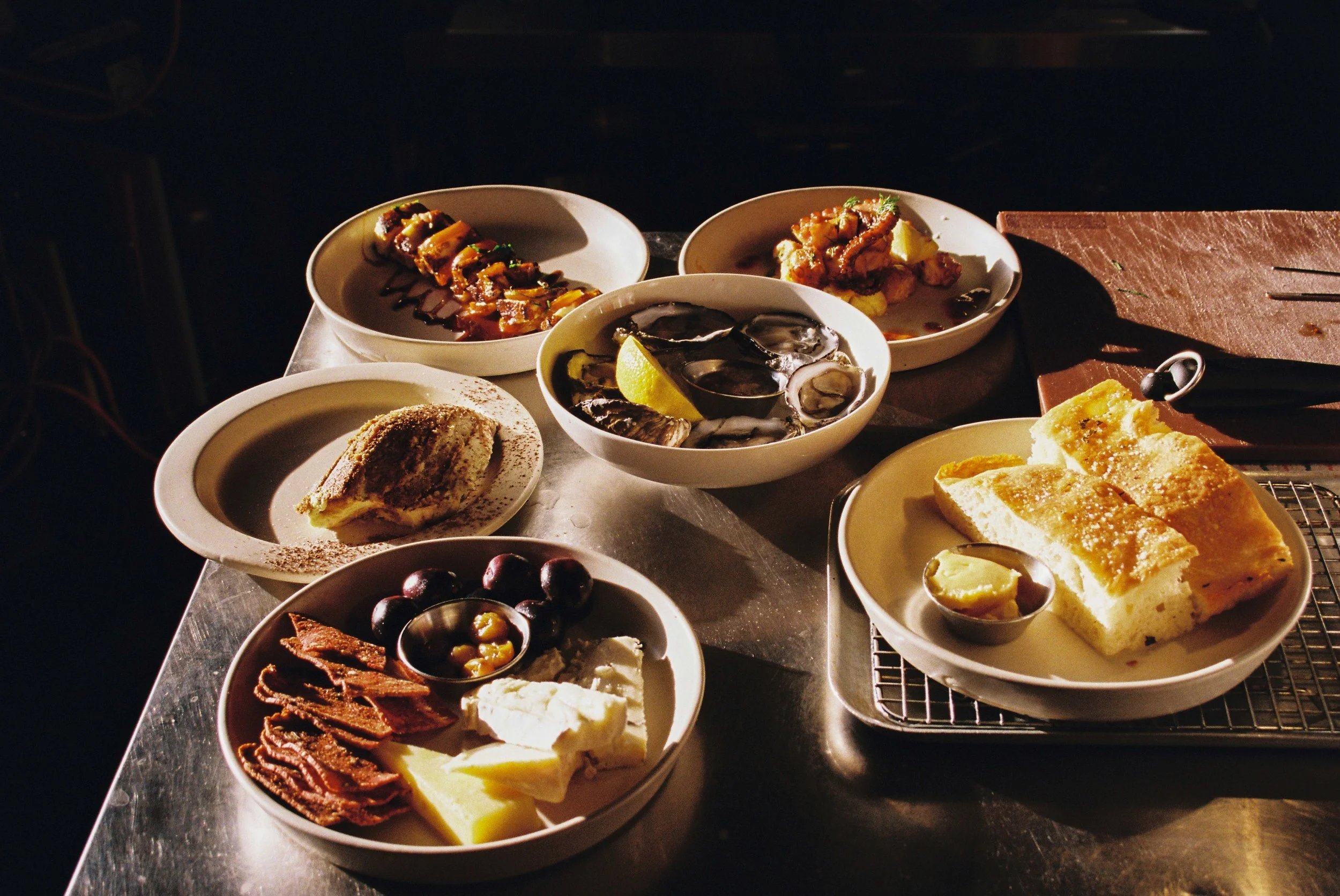 Assorted dishes including seafood, bread, cheese, olives, and desserts on a table in warm lighting. Gold Coast Food Photography by Joey Bailey