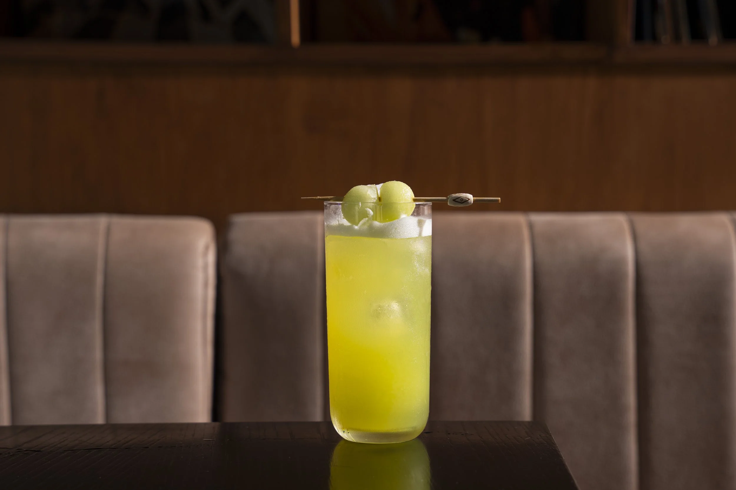 A tall glass of bright green cocktail with ice, garnished with melon balls on a skewer, placed on a dark table in front of a beige cushioned booth. Gold Coast | Byron Bay Food Photography by Joey Bailey
