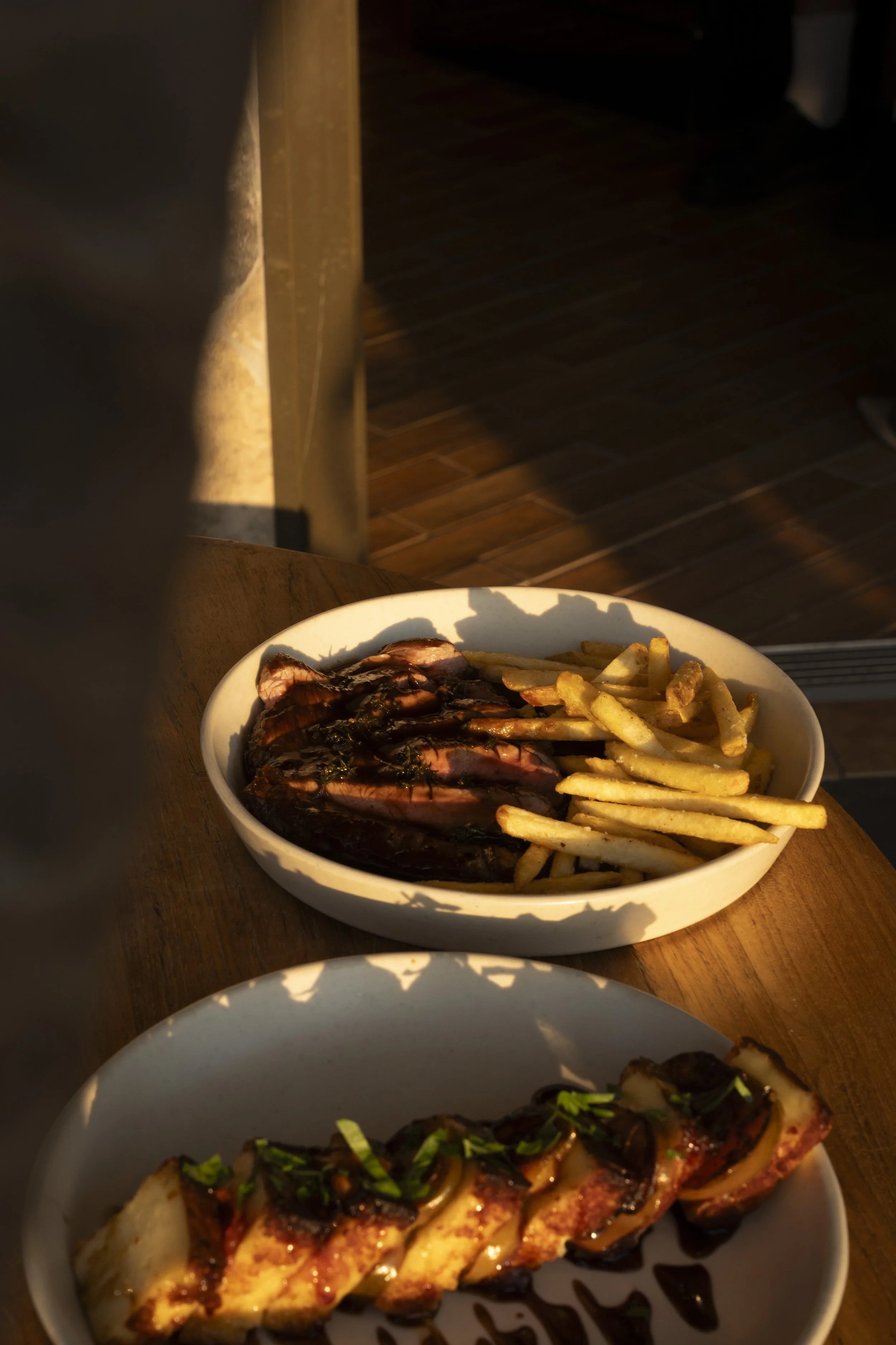 A white bowl with grilled steak topped with sauce, alongside French fries on a wooden table, with warm lighting. Gold Coast Food Photography by Joey Bailey