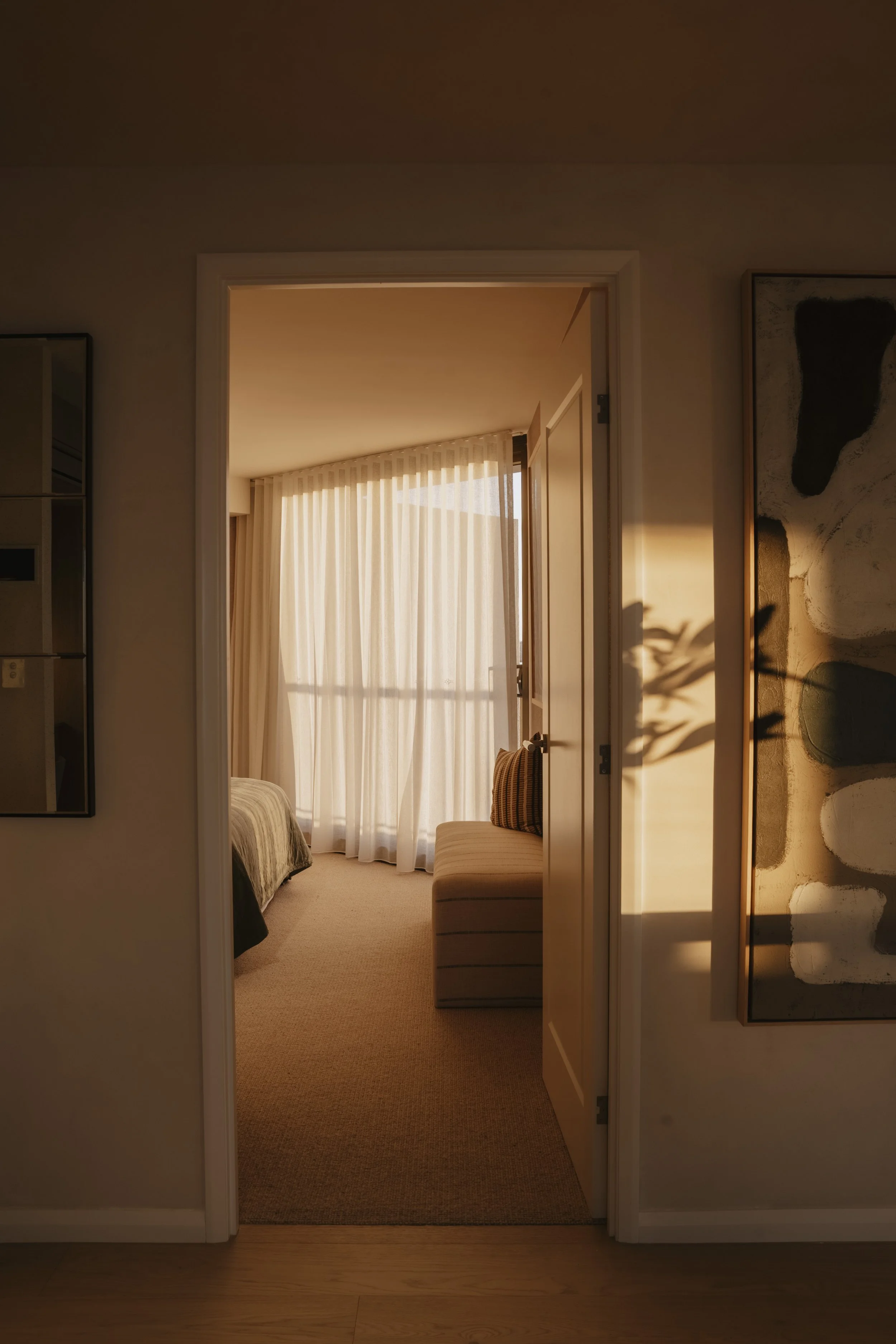 A cozy bedroom with sunlight streaming through sheer curtains, a bed partially visible on the left, and a beige striped bench or ottoman in the center. Gold Coast Architecture photography by Joey Bailey