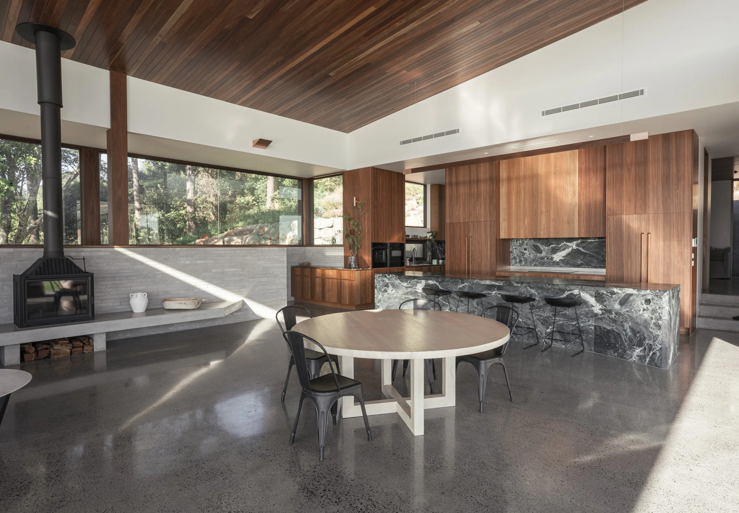 Modern living room with large windows, wood and marble accents, a round dining table with black chairs, a wood-paneled ceiling, and a stone fireplace. Cape House, Suffolk Park – architectural photography