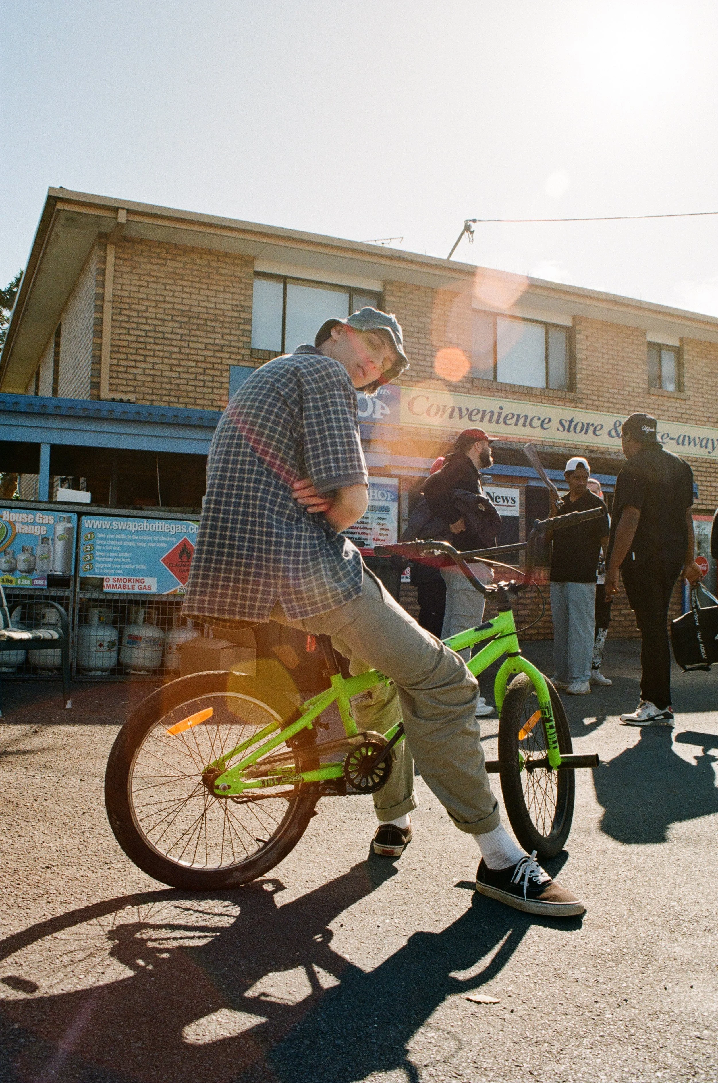 Person on green bicycle in front of convenience store, group of people in the background, sunlight lens flare