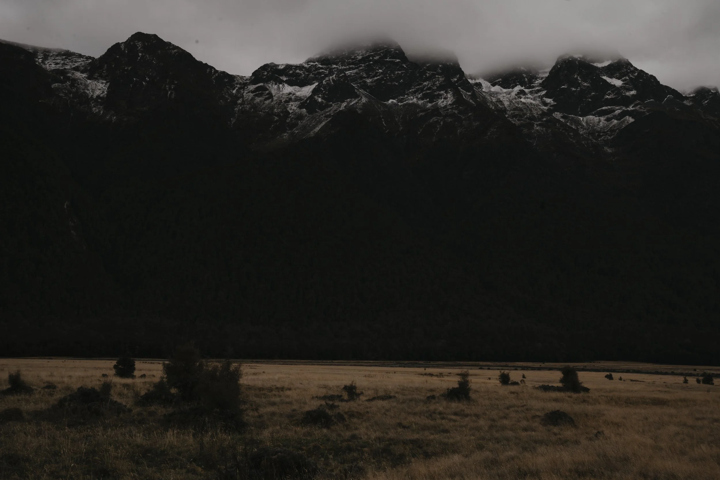 Dark mountain landscape with a grassy field in the foreground, misty clouds covering the mountain peaks.
