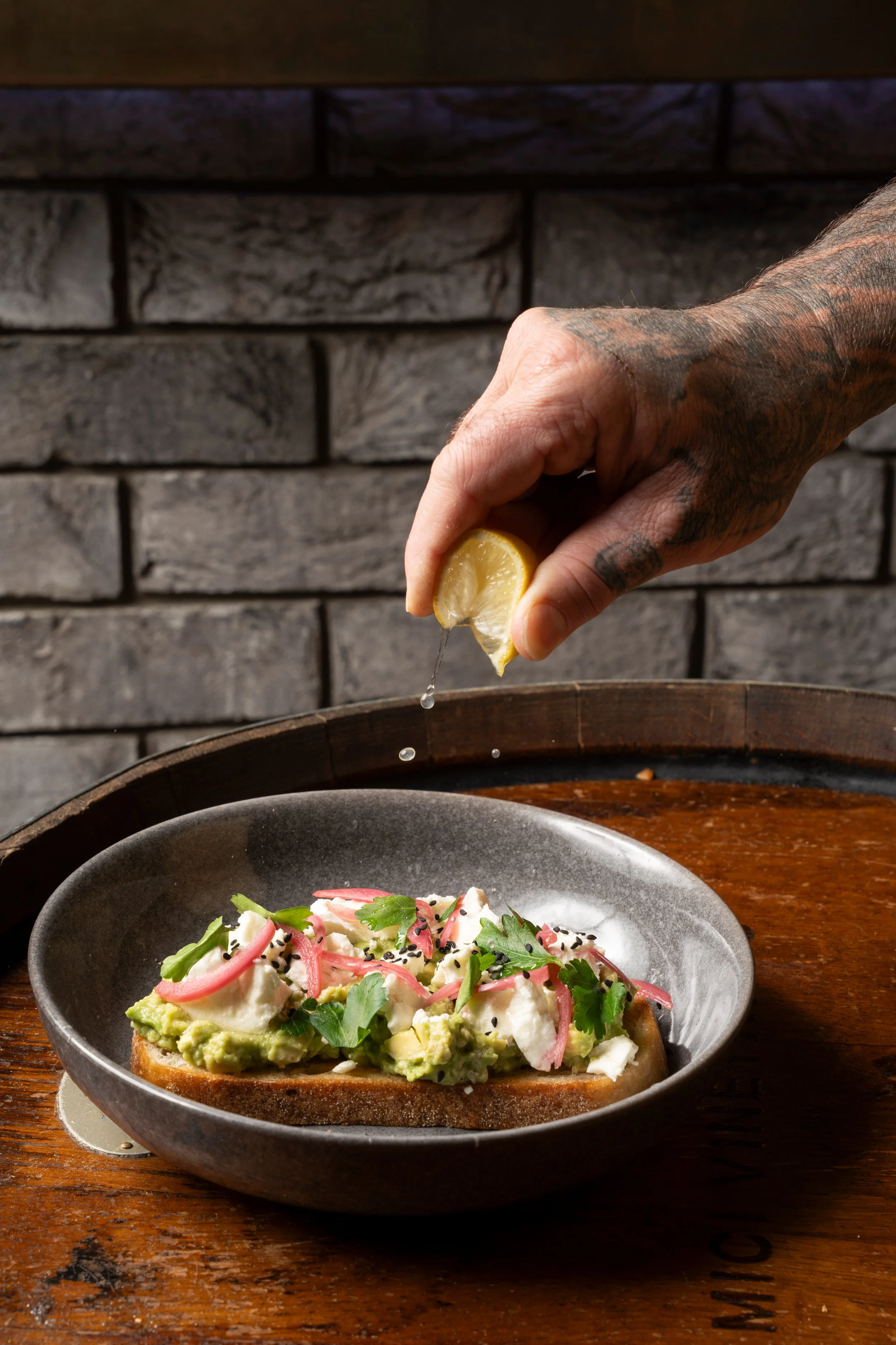 Hand squeezing lemon over an avocado toast topped with radish slices, herbs, and cream, on a black plate placed on a wooden table. Gold Coast food photography by Joey Bailey
