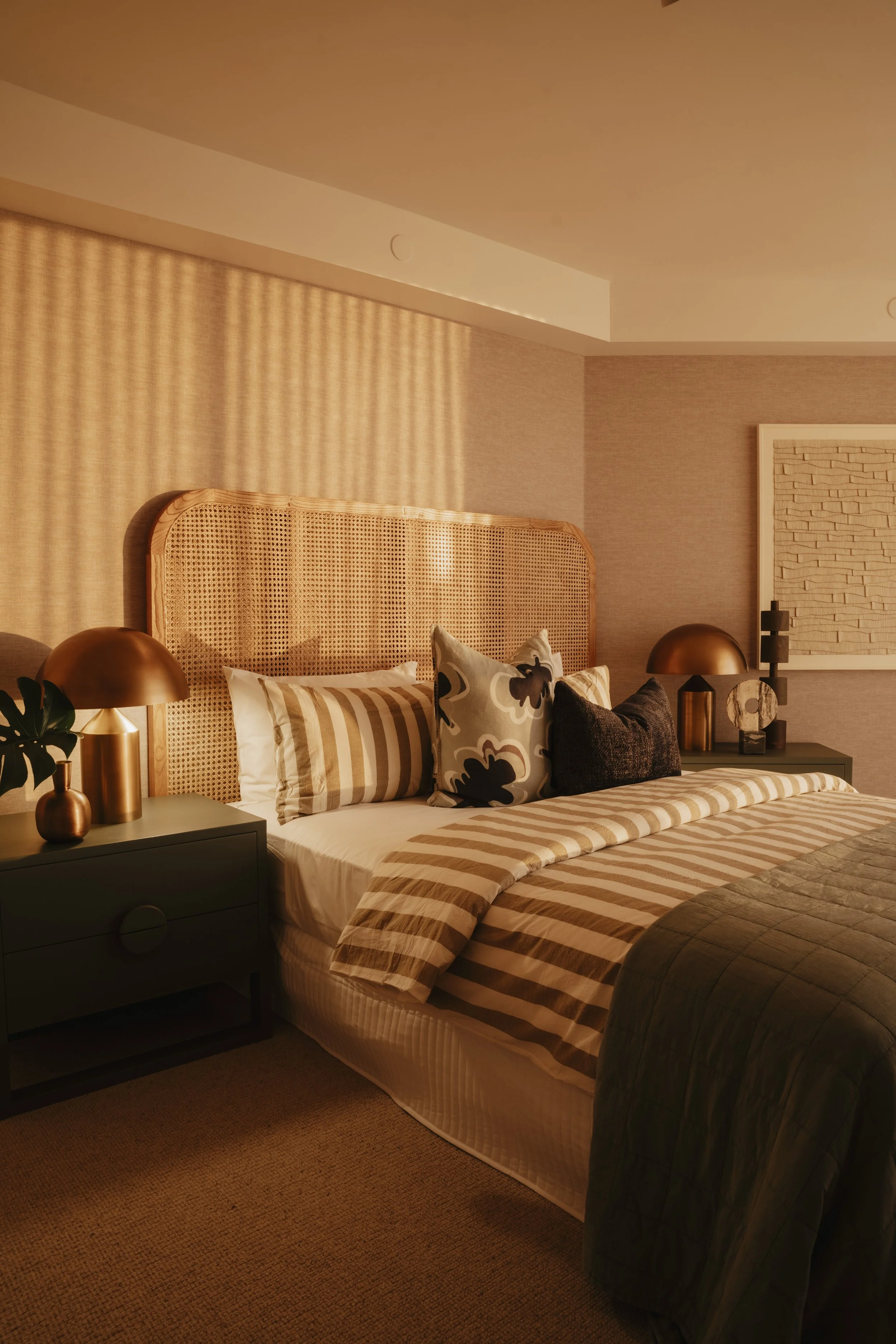 A cozy bedroom with a rattan headboard, striped bed linens, multiple decorative pillows, matching bedside tables with gold lamps, and textured wall art. Gold Coast Architecture photography Joey Bailey