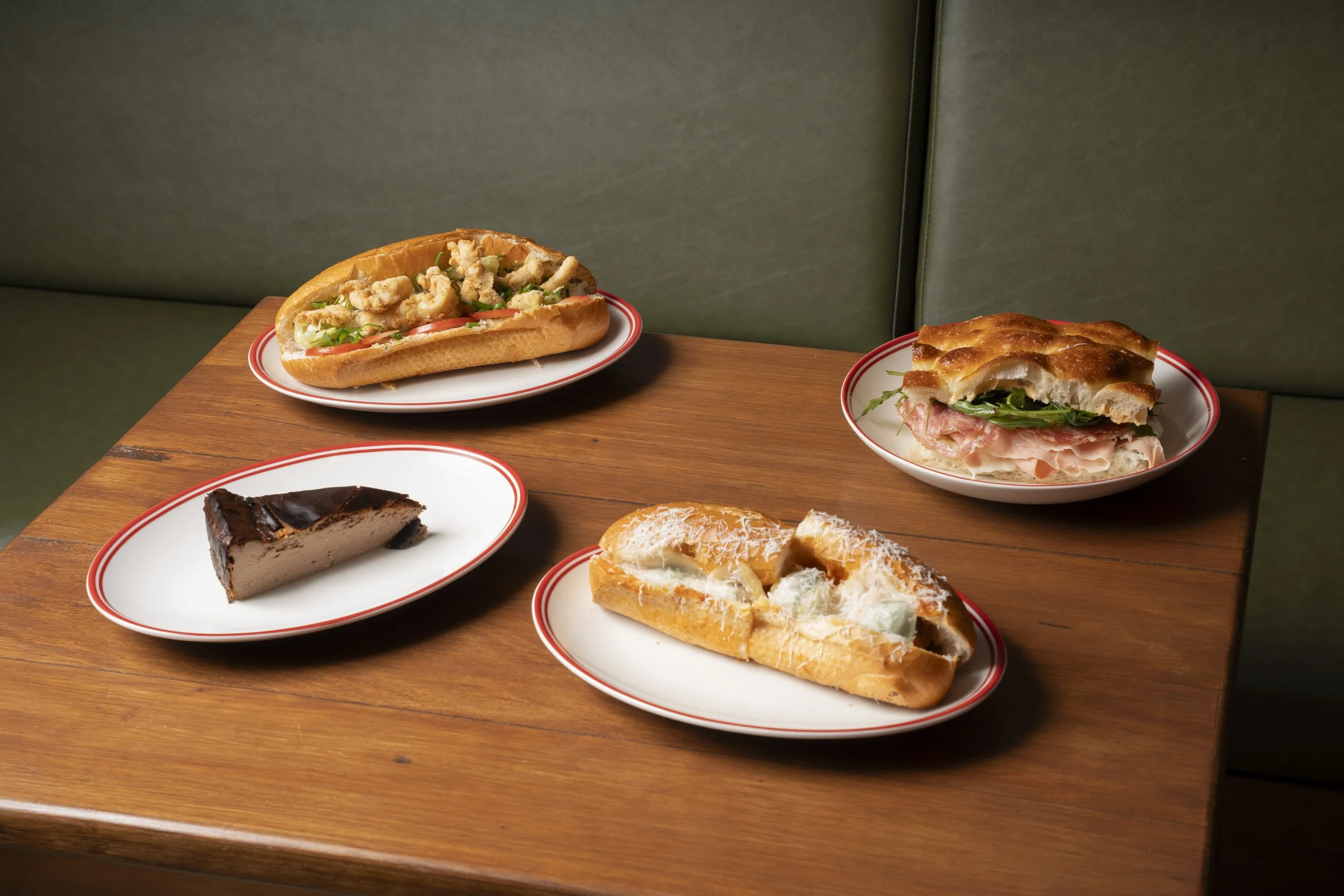 Assorted desserts and sandwiches on plates on a wooden table with a green cushioned background.