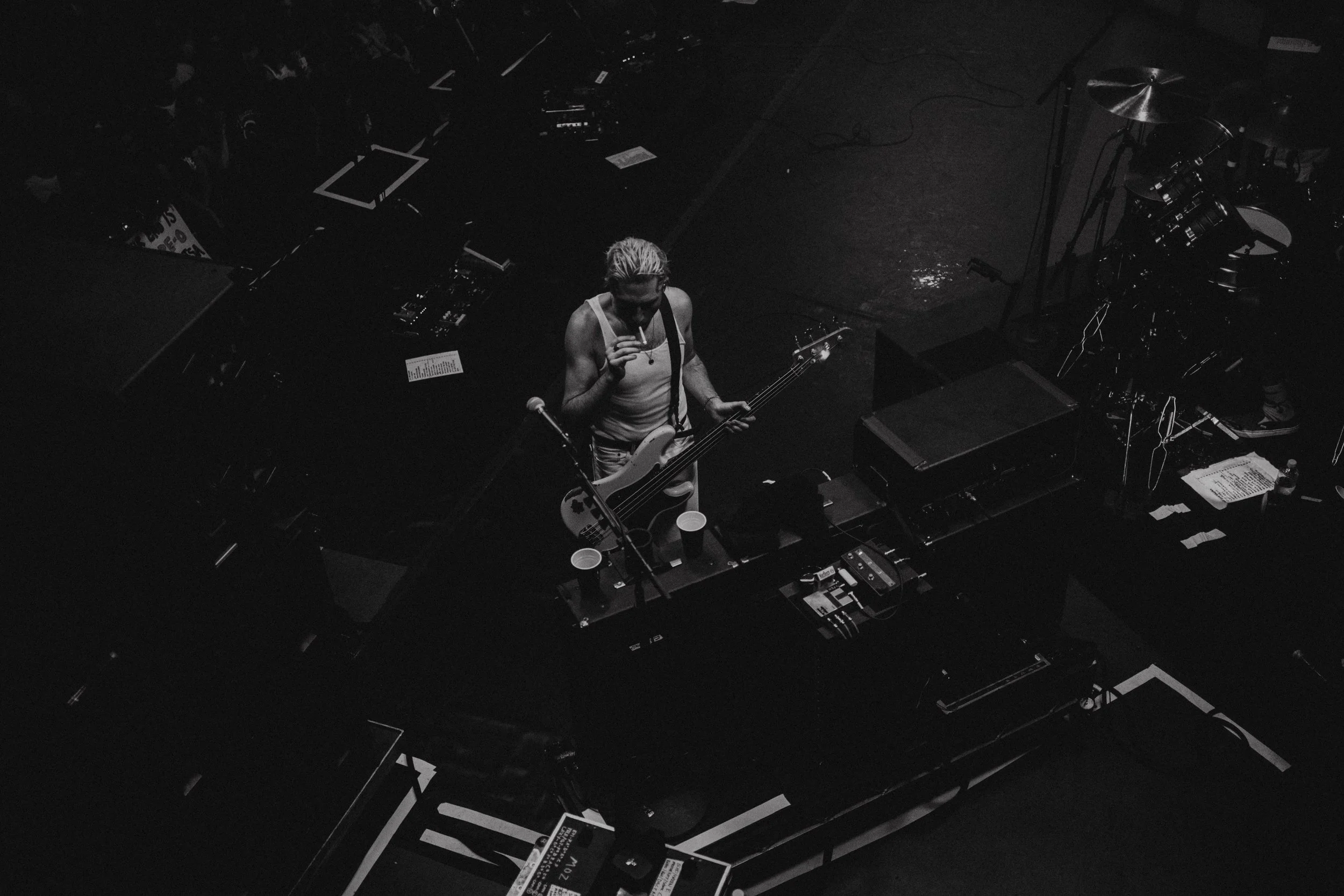 Black and white photo of a musician on stage holding a bass guitar near a microphone stand, surrounded by stage equipment and a drum set.