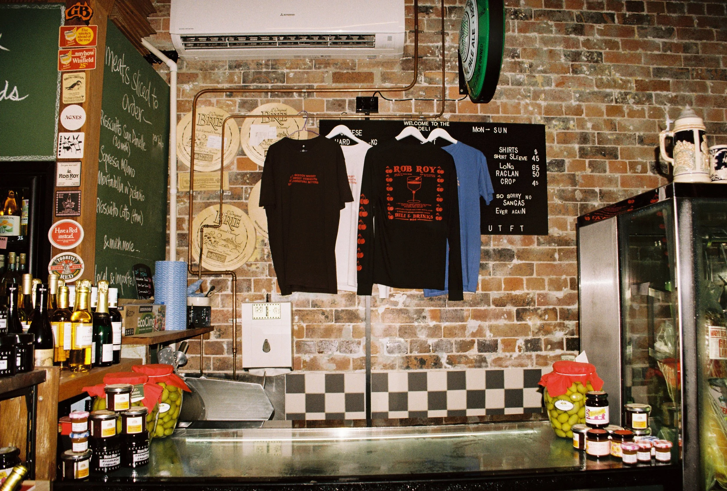 Interior of a deli or bar featuring a brick wall with hanging T-shirts and a blackboard menu. The T-shirts advertise Rob Roy and feature red graphics. Tweed heads marketing and brand photography 