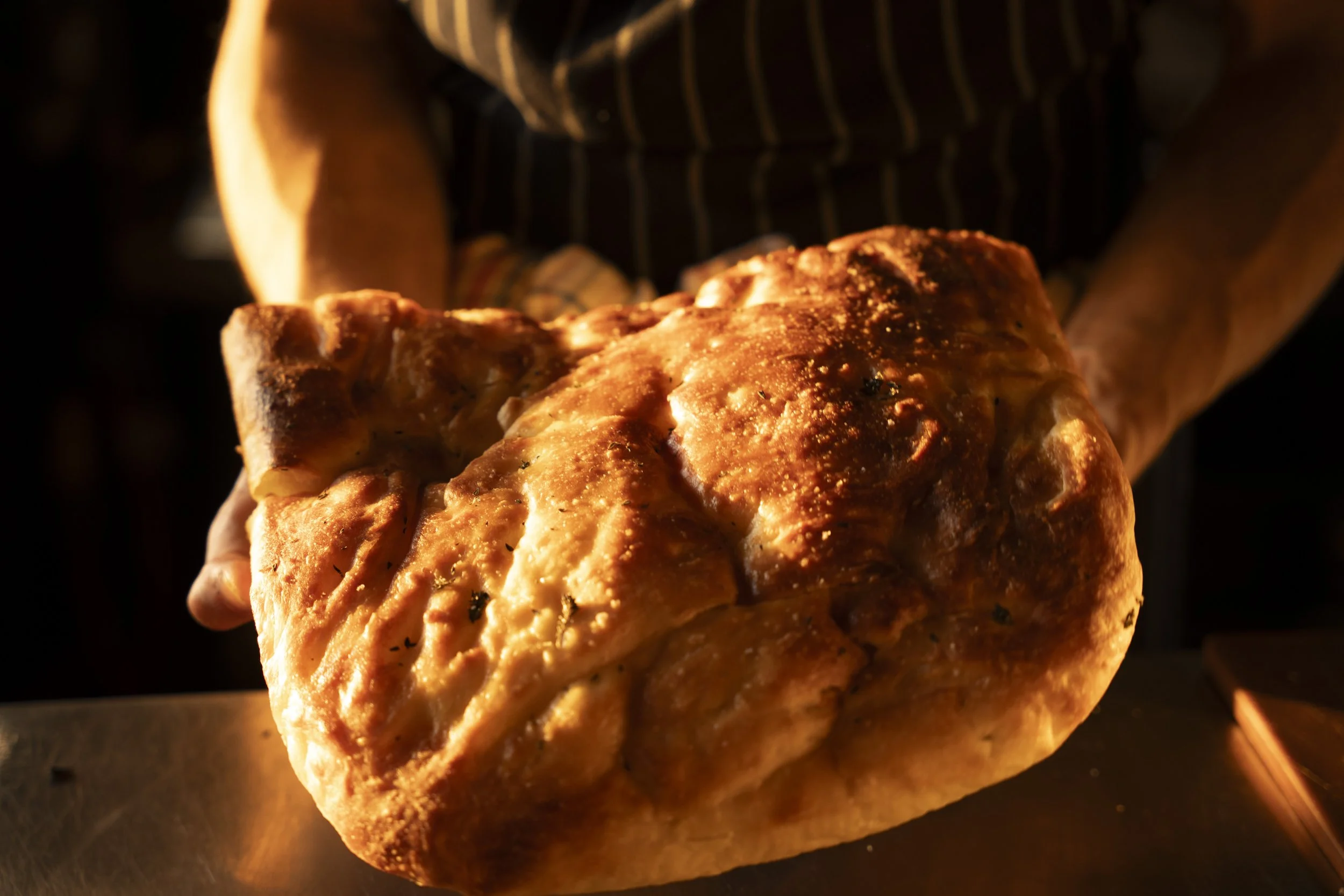 A person holding a freshly baked, golden brown loaf of bread, with a crispy crust, in a dark setting. Gold Coast Food Photography by Joey Bailey