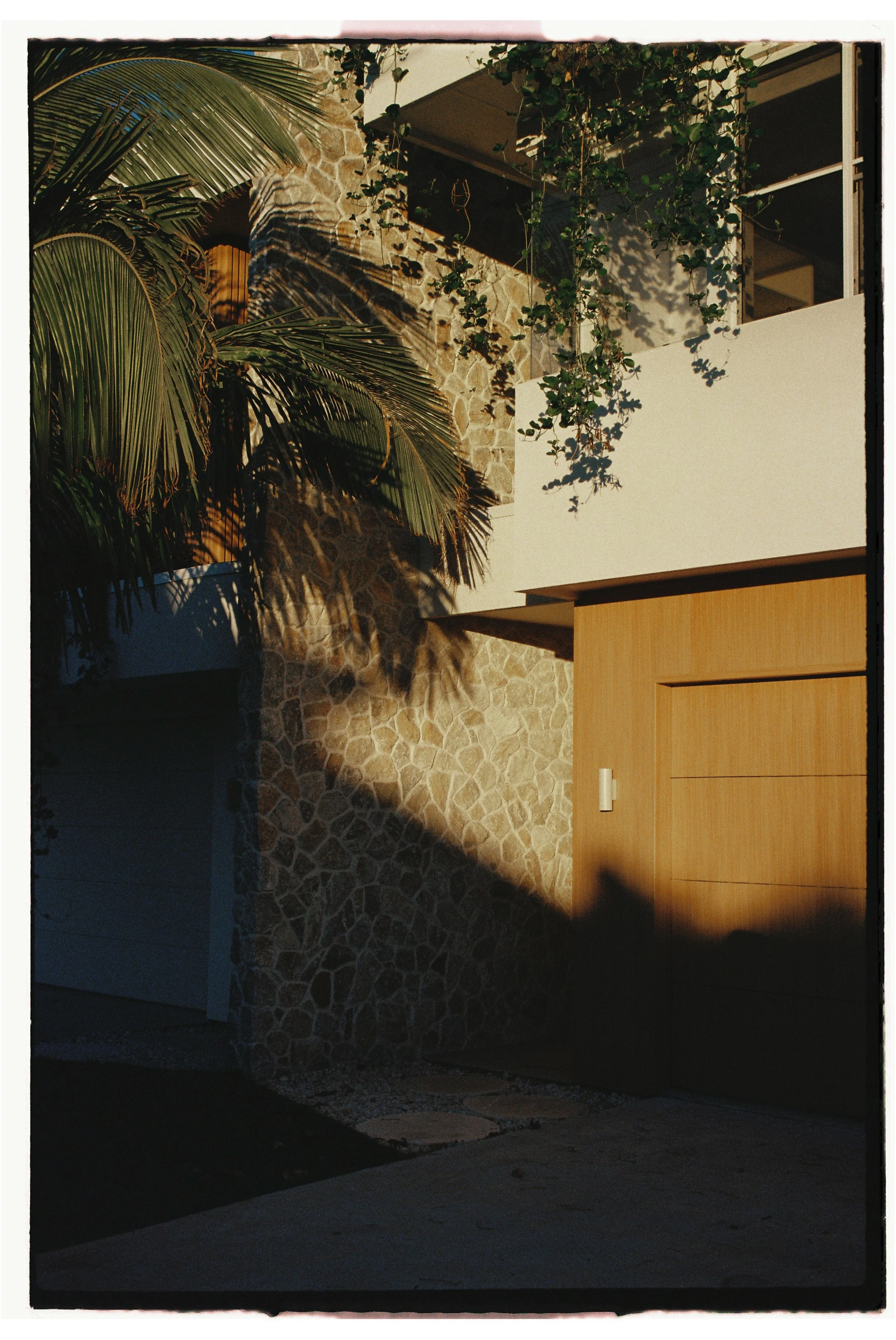 A modern house exterior with a stone wall, wooden garage door, and lush green plants casting shadows at sunset. Architecture photography Gold Coast by Joey Bailey