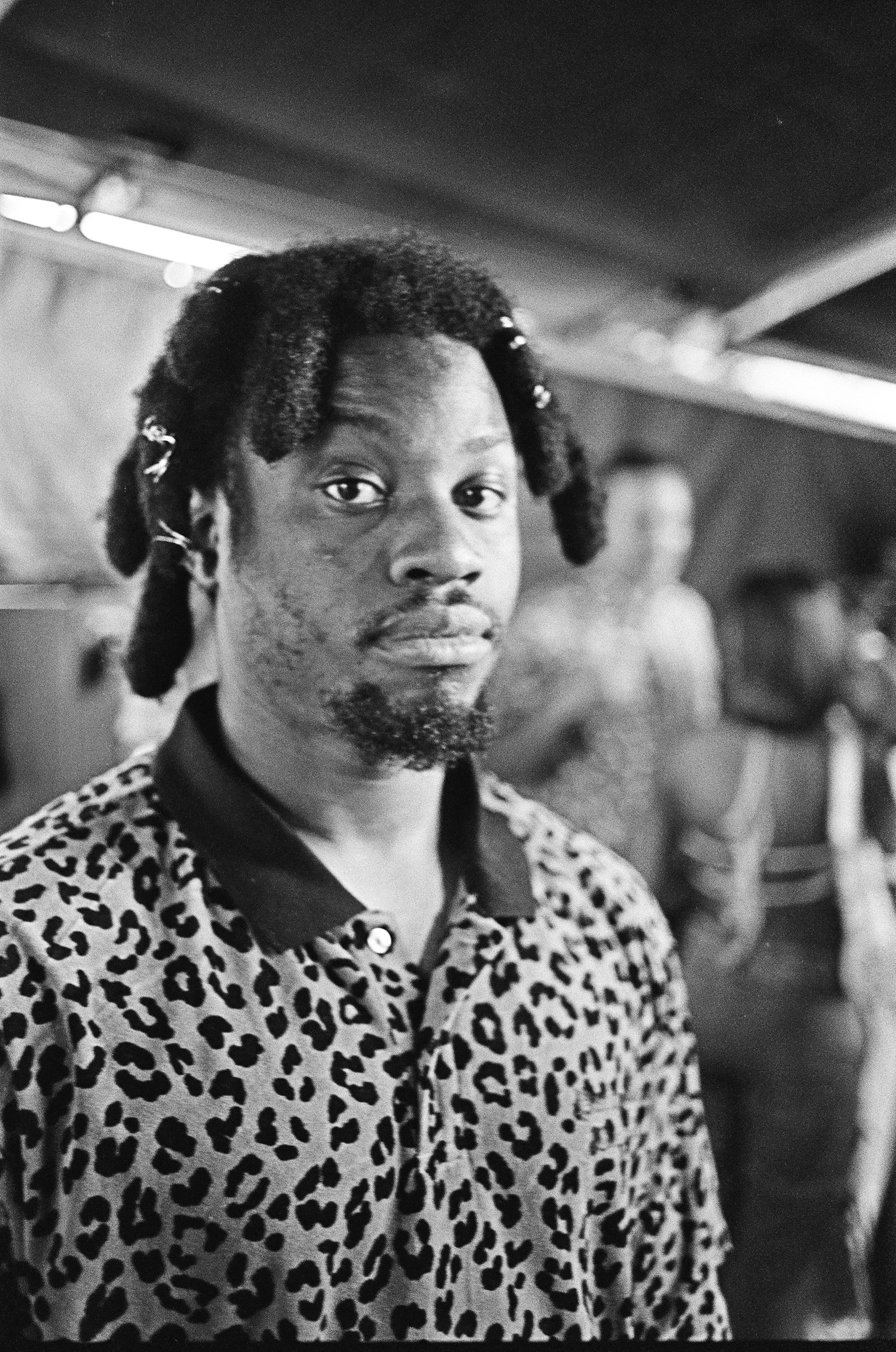 Black and white photo of a person with short dreads, wearing a leopard print shirt in a room with people in the background.