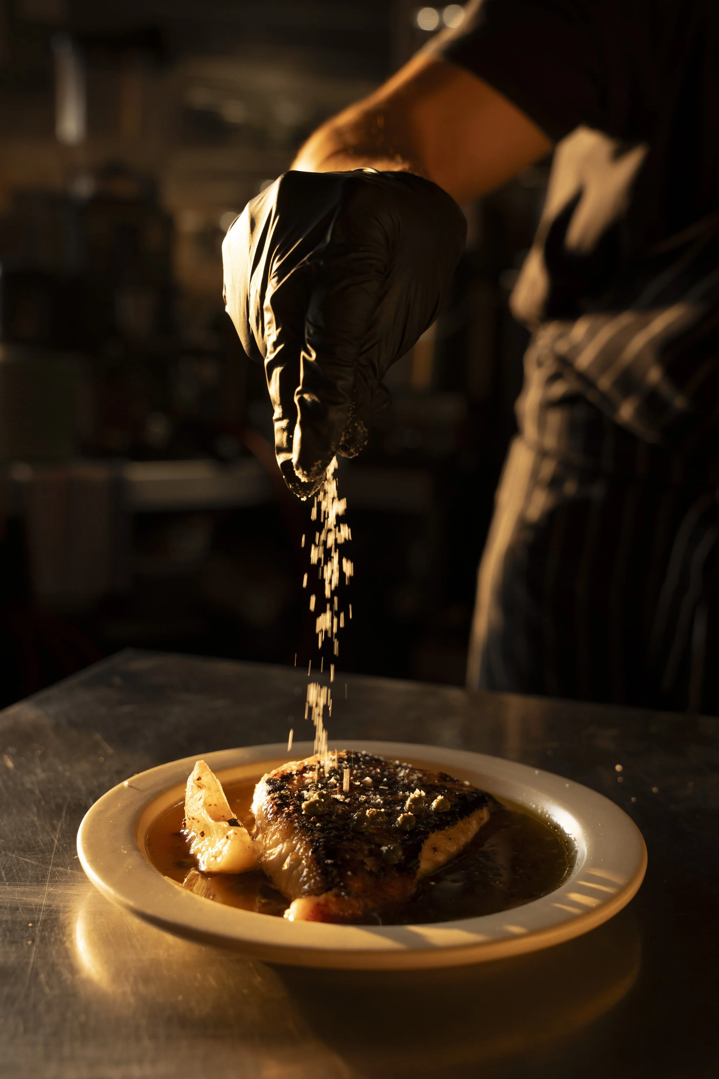 A chef seasoning a cooked fish filet with salt over a plate of fish with sauce in a dimly lit kitchen. Gold Coast Food Photography by Joey Bailey