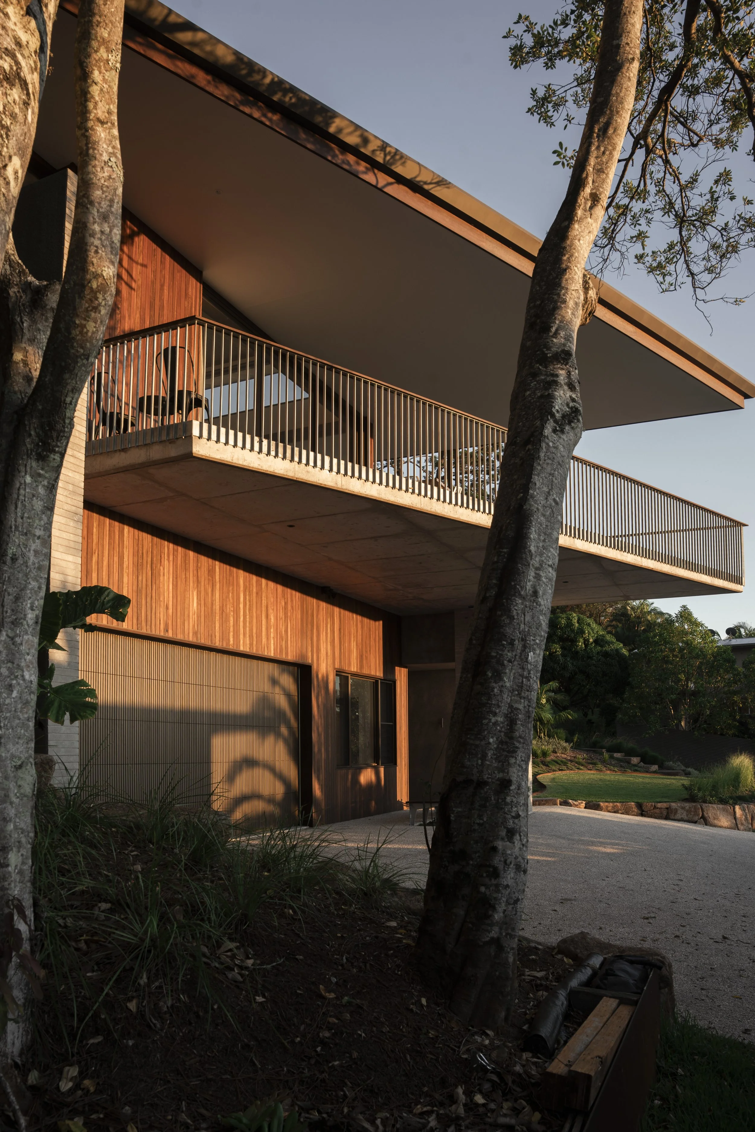 A modern house with wooden siding and large glass windows, featuring a spacious balcony with outdoor furniture, surrounded by trees and greenery during sunset. Cape House, Suffolk Park – architectural photography