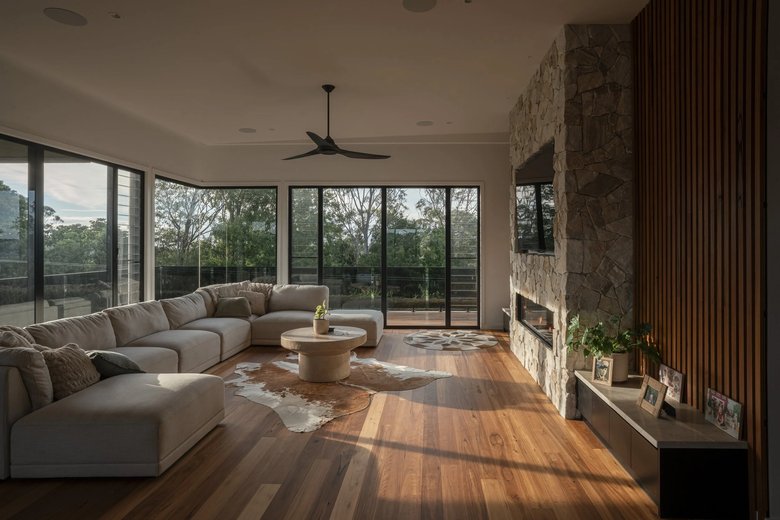 Modern living room with large beige sectional sofa, round coffee table, cowhide rug, stone fireplace, wooden floor, and expansive glass sliding doors showing trees outside. Architecture photography by Joey Bailey Gold Coast