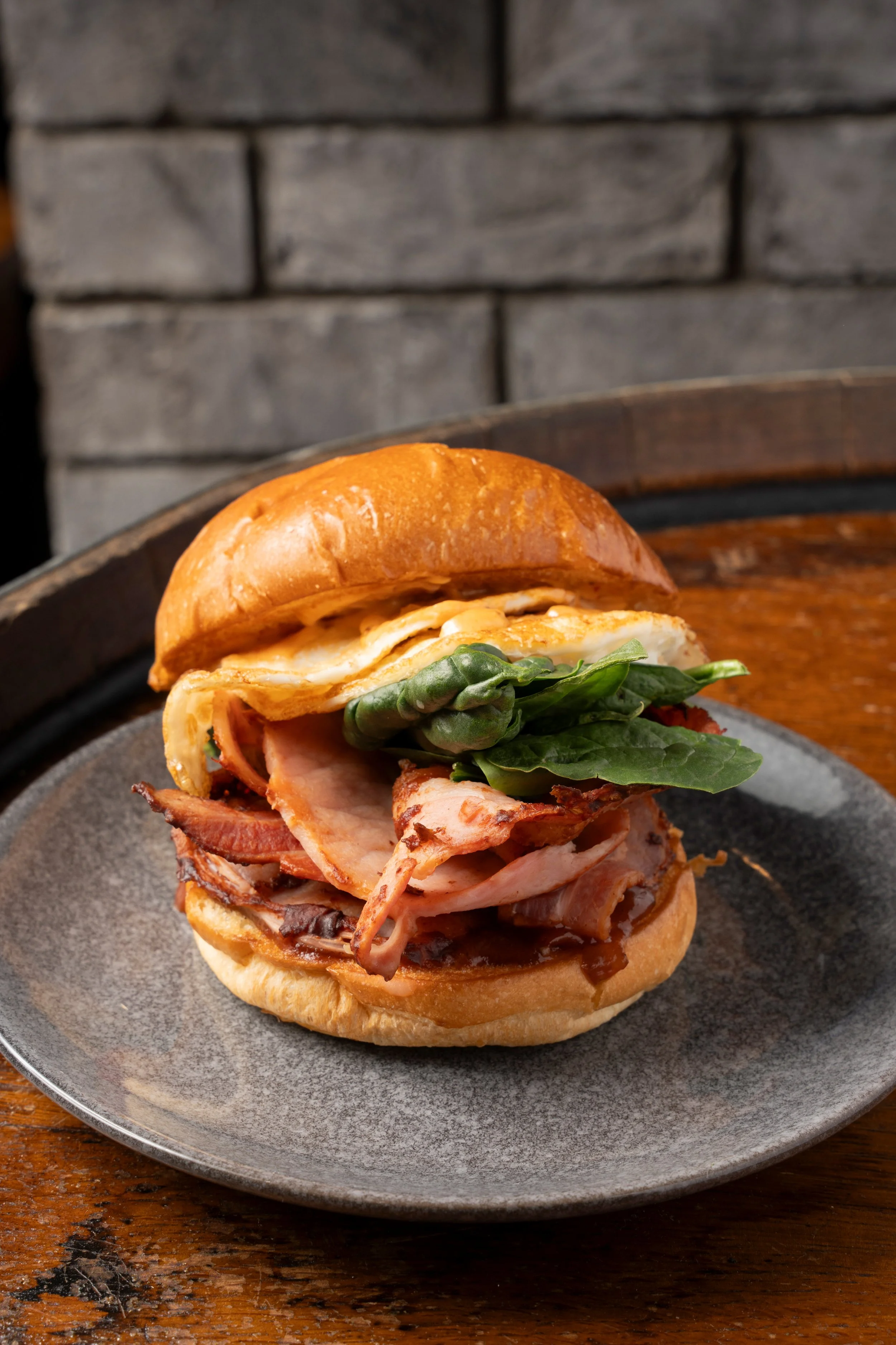 Sandwich with bacon, ham, lettuce, fried egg, on toasted bun, on a gray plate with rustic wooden table and stone wall background. Gold Coast food photography by Joey Bailey